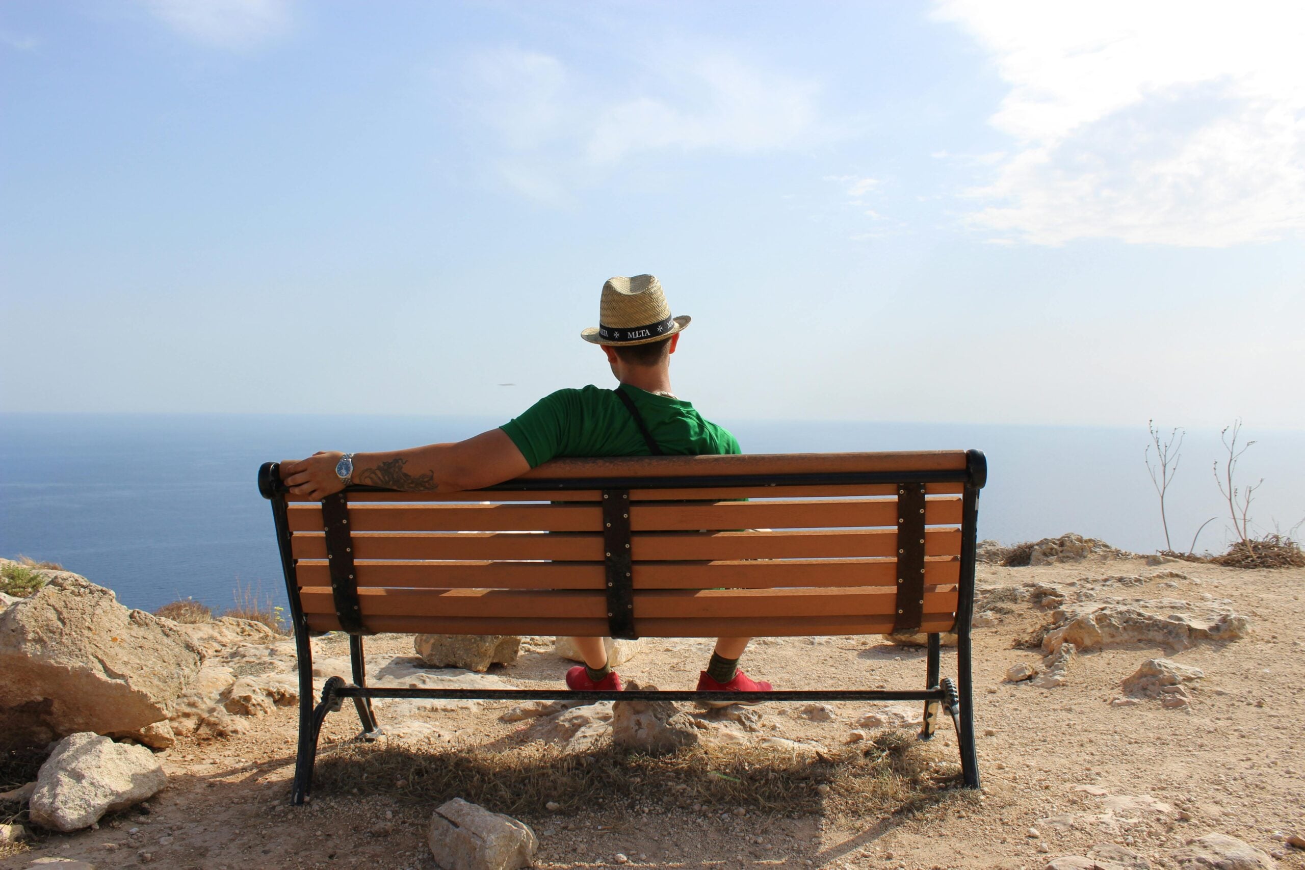 A person wearing a hat and green shirt sits on a wooden bench, facing the ocean and clear sky, enjoying a scenic, rocky coastal view.