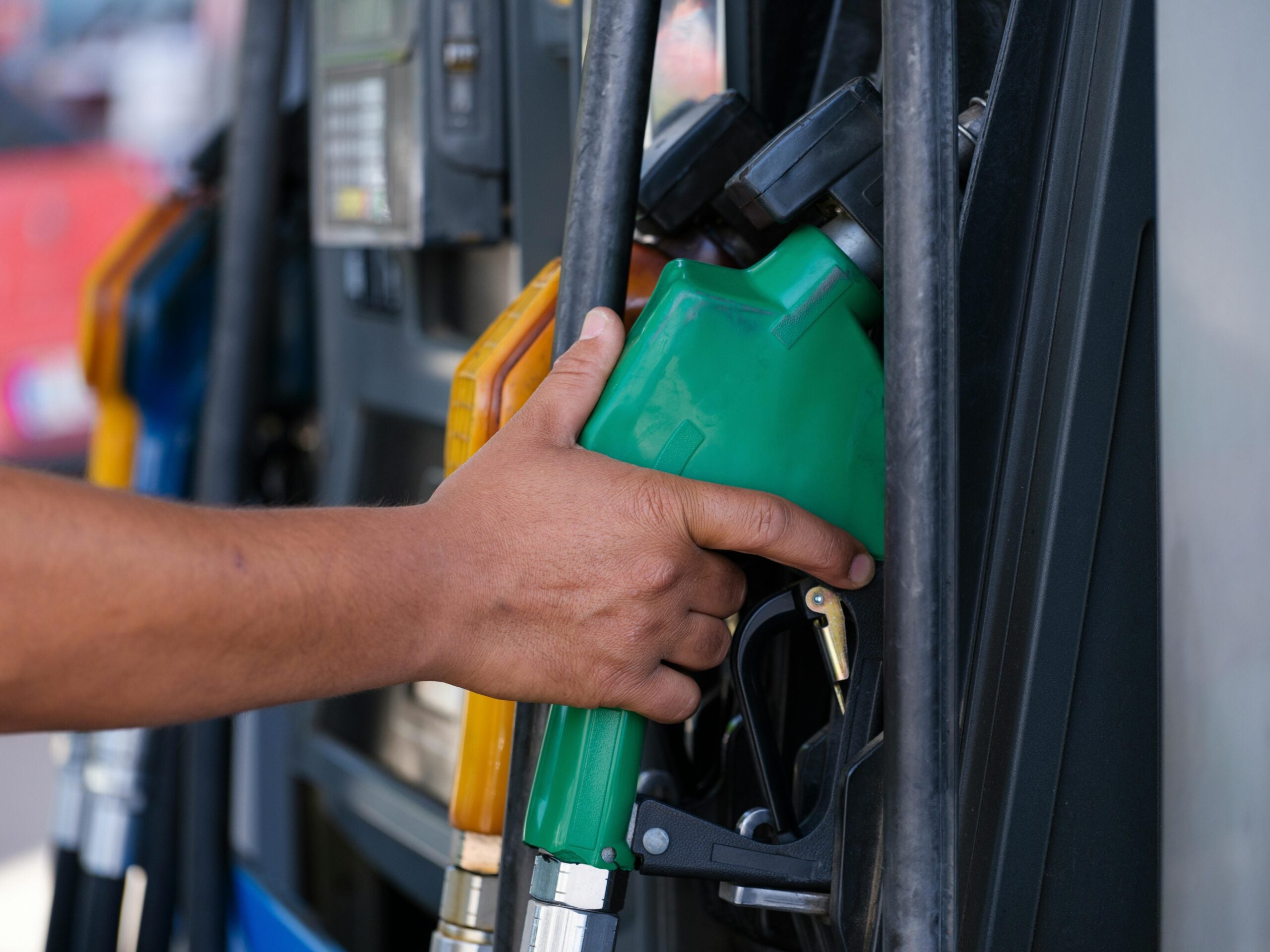 A person’s hand reaches for a green fuel pump nozzle at a gas station, with other fuel nozzles visible in the background.