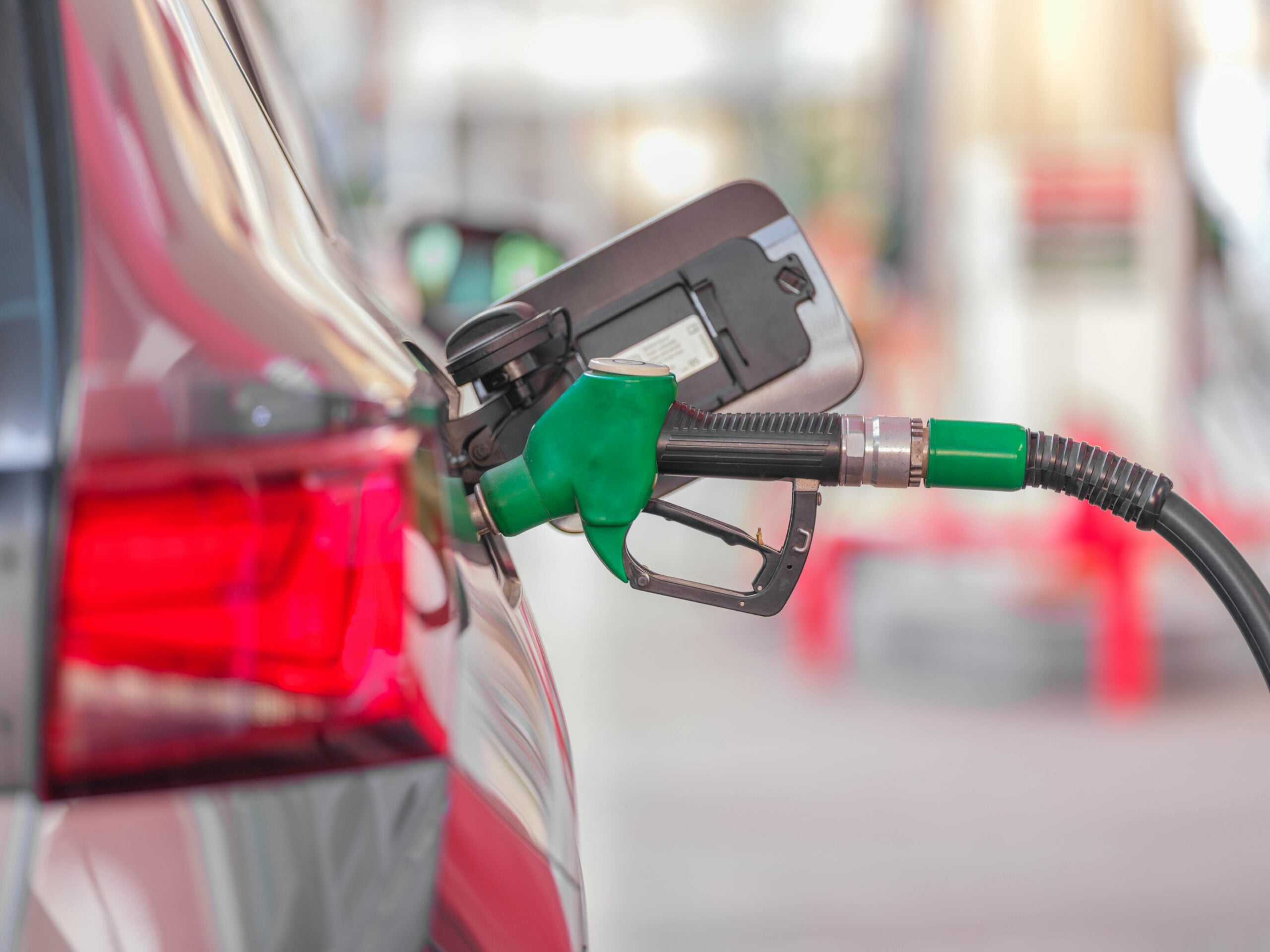 A green fuel nozzle is inserted into the fuel tank of a silver car at a gas station, with the car’s taillight visible and the background blurred.