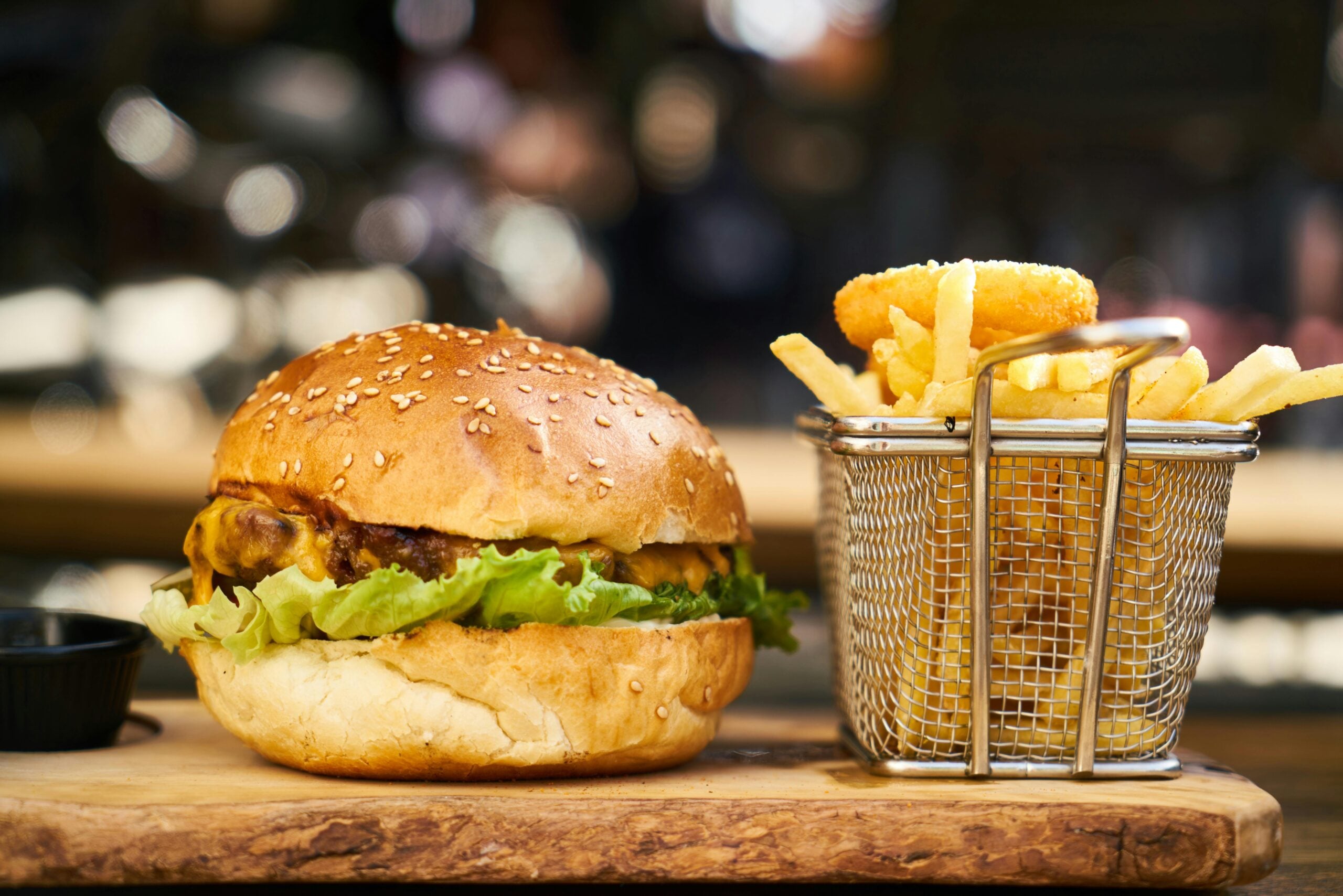 A close-up of a cheeseburger with lettuce on a wooden board, next to a metal basket filled with French fries and onion rings. A small black sauce cup is placed to the side.