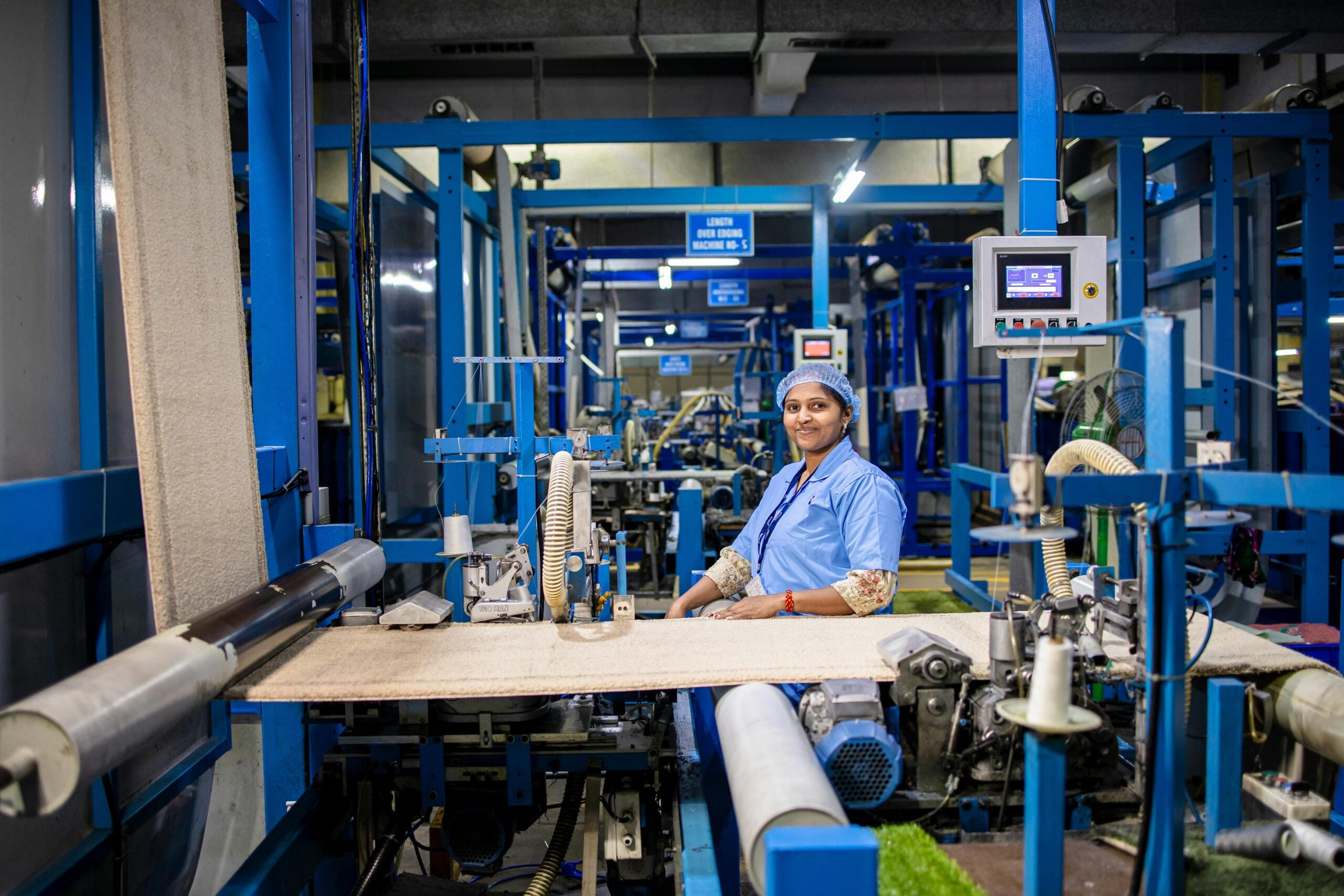 A woman in blue work attire stands smiling at a workstation in a modern textile factory, surrounded by industrial machinery and fabric rolls.