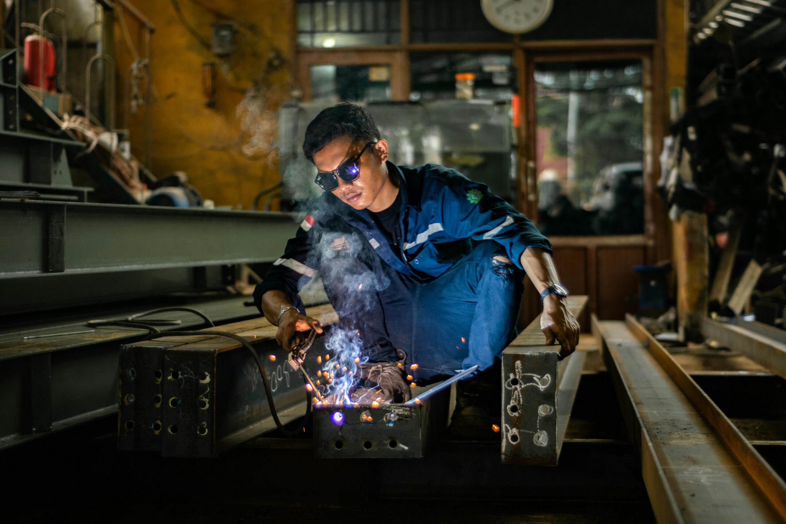 A person wearing protective glasses and work clothes welds metal beams in an industrial workshop, surrounded by tools and equipment, with sparks and smoke visible from the welding process.