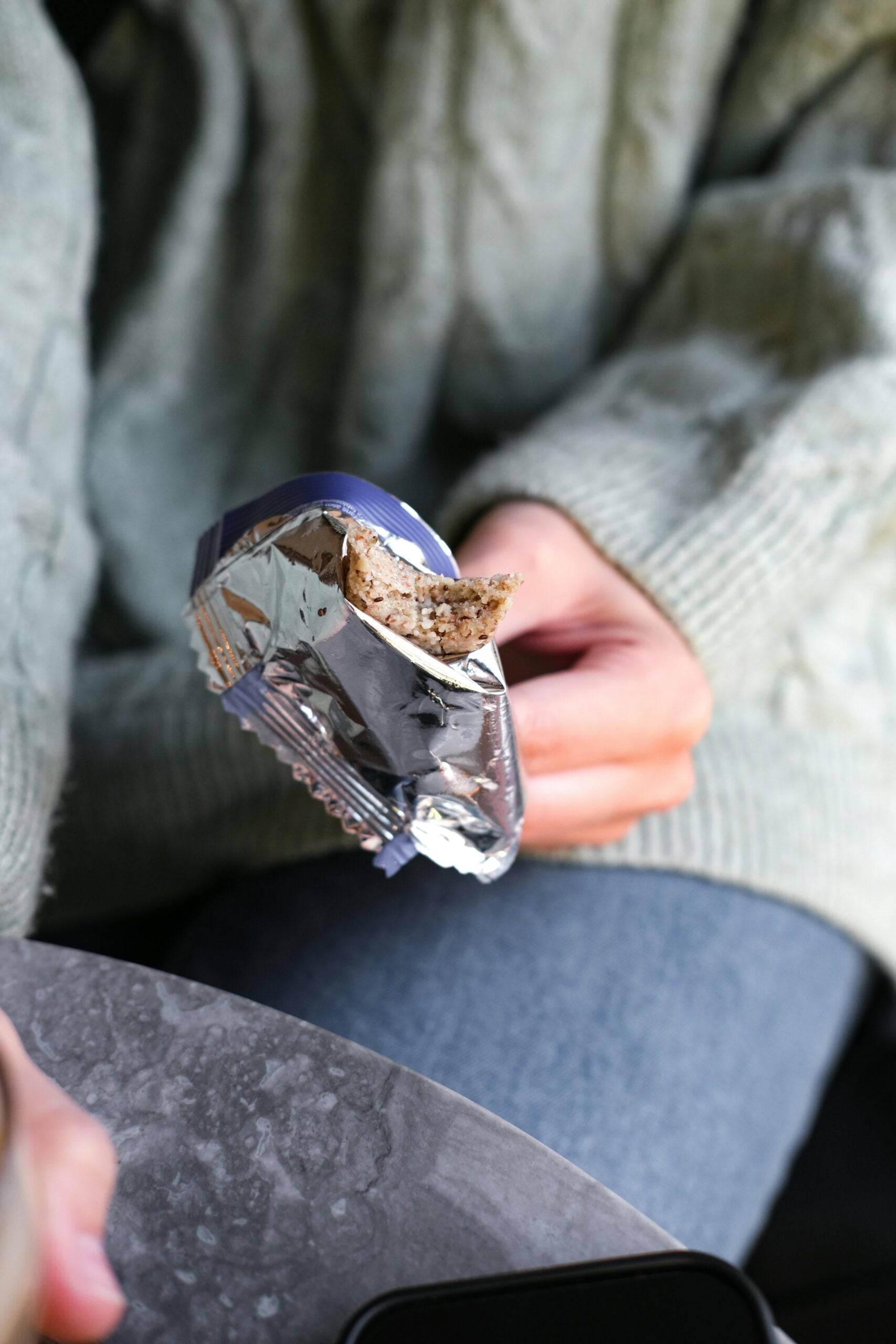 A person wearing a light green sweater and blue jeans holds a partially unwrapped granola bar with a bite taken out of it, sitting next to a round gray table.
