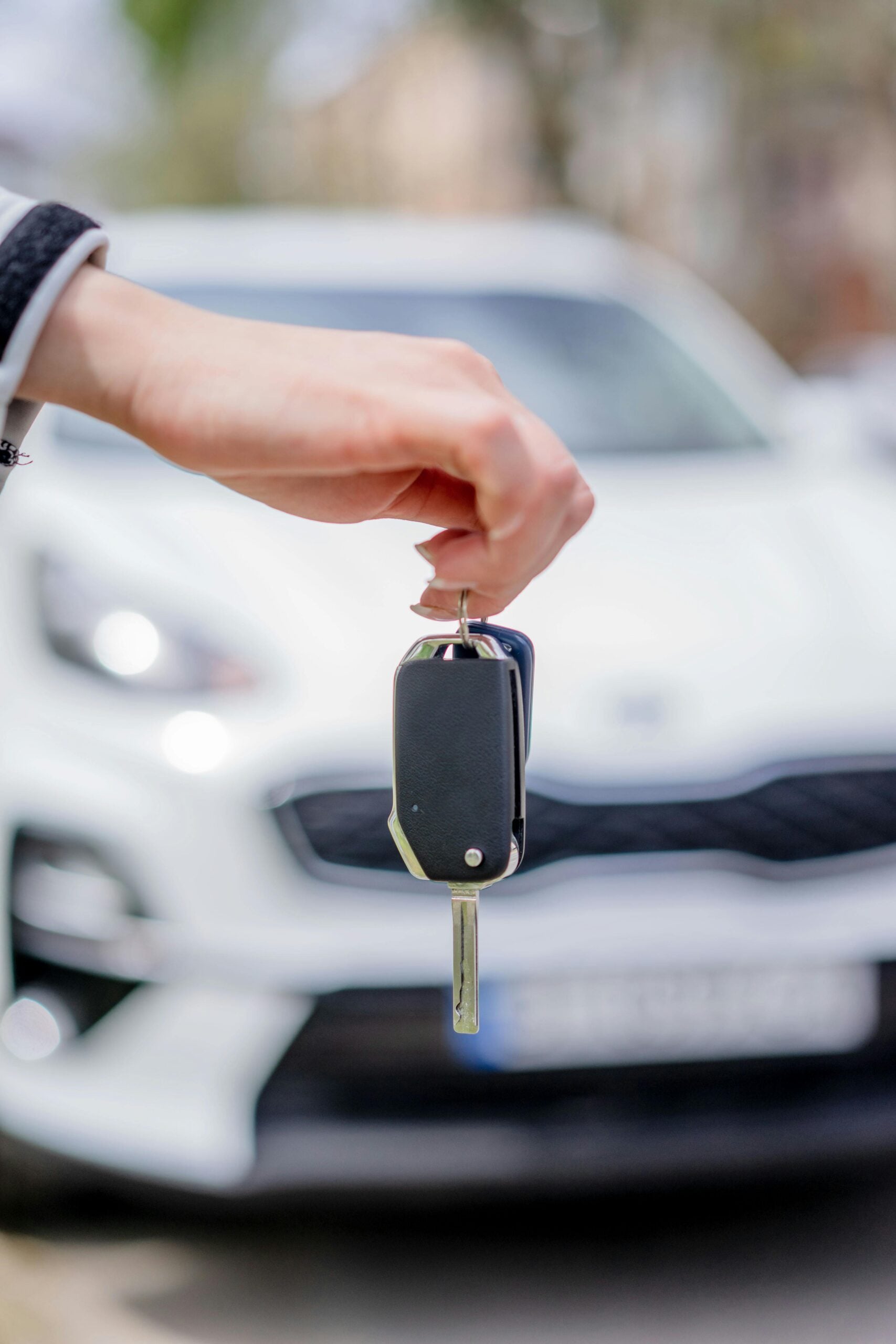 A hand holds a car key in front of a blurred white car, suggesting themes of driving, car ownership, or vehicle purchase.