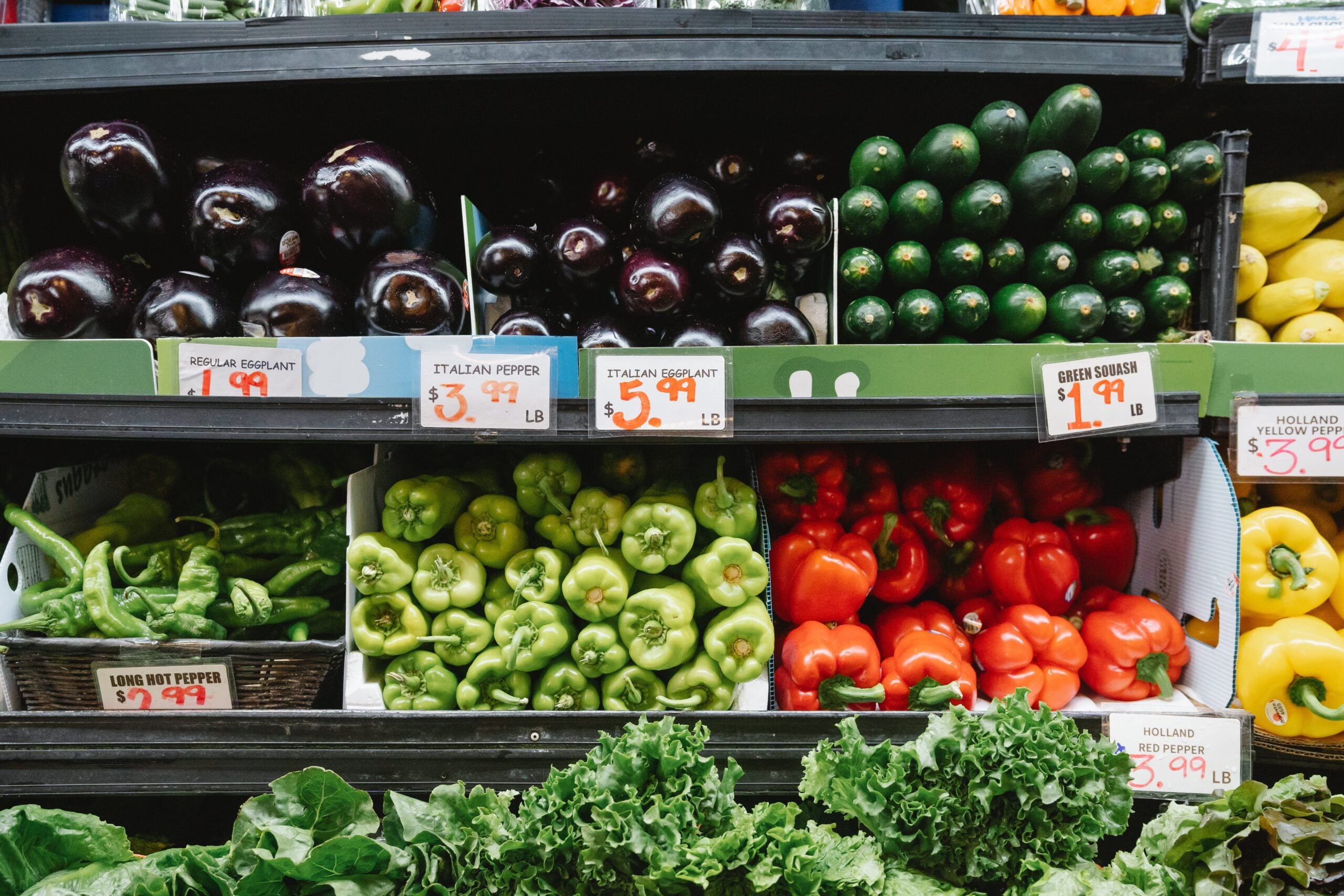 A grocery store produce display with eggplants, zucchini, green and red bell peppers, hot peppers, yellow bell peppers, and leafy greens, all labeled with price tags.