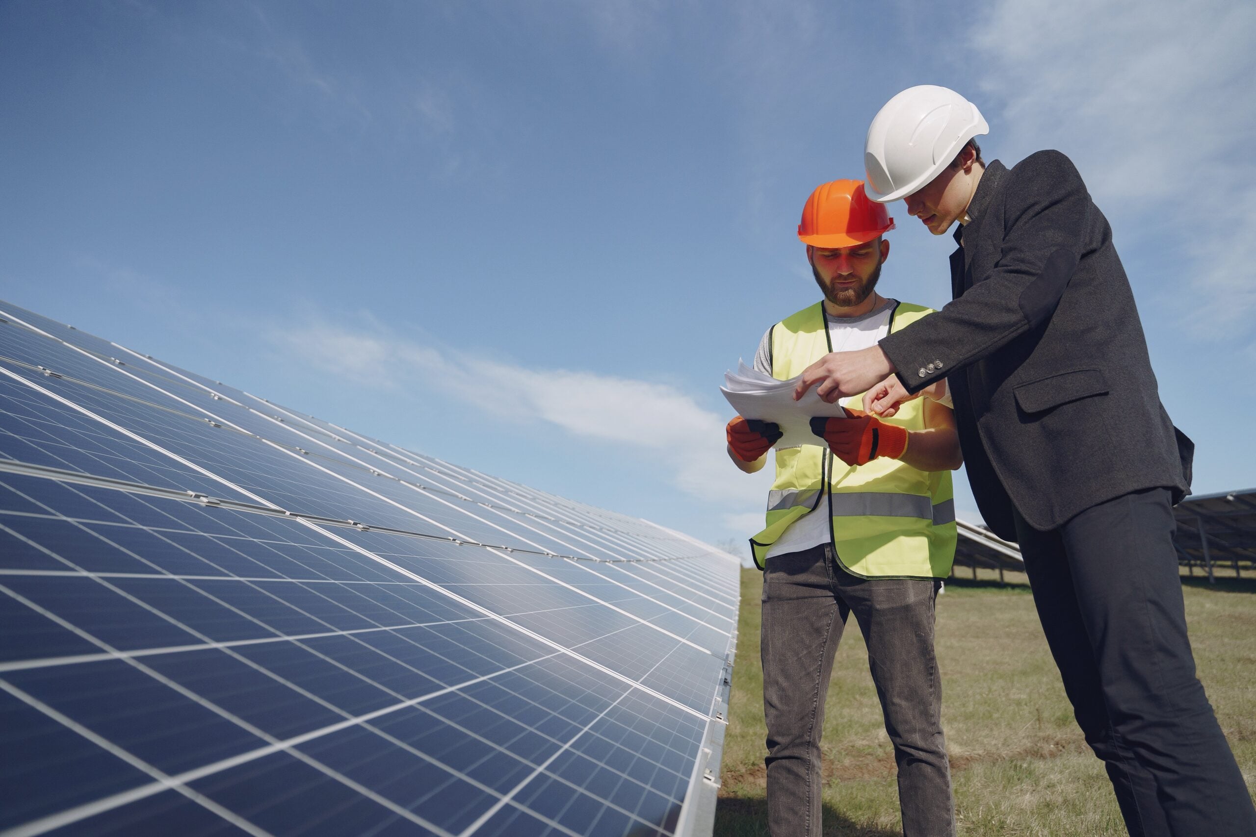 Two men wearing safety helmets and vests stand beside solar panels outdoors, reviewing documents together under a clear blue sky.