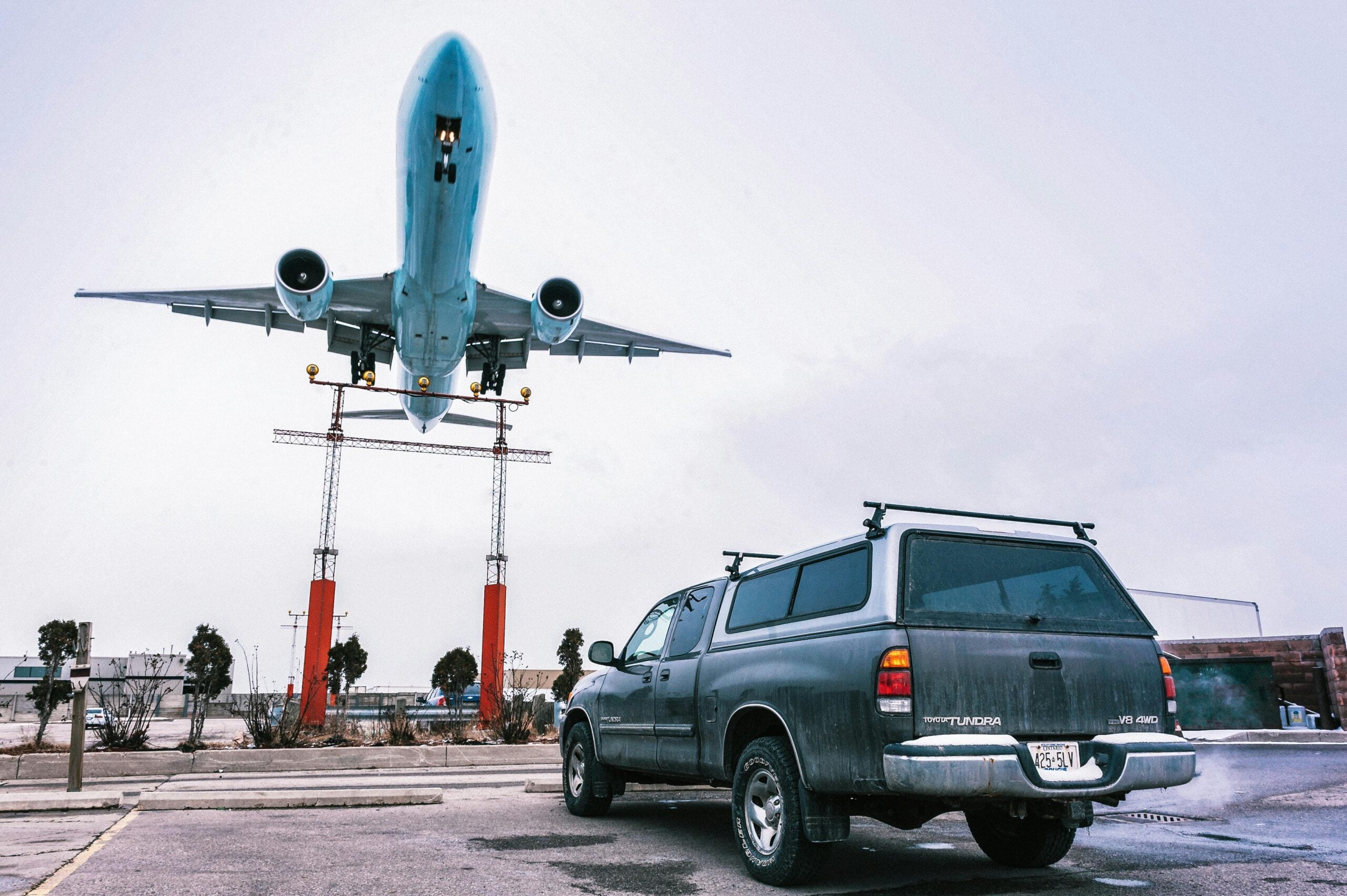 A large airplane flies low overhead, about to land, above a gray pickup truck parked in a lot near the runway. The scene is cloudy with some buildings, trees, and landing lights in the background.