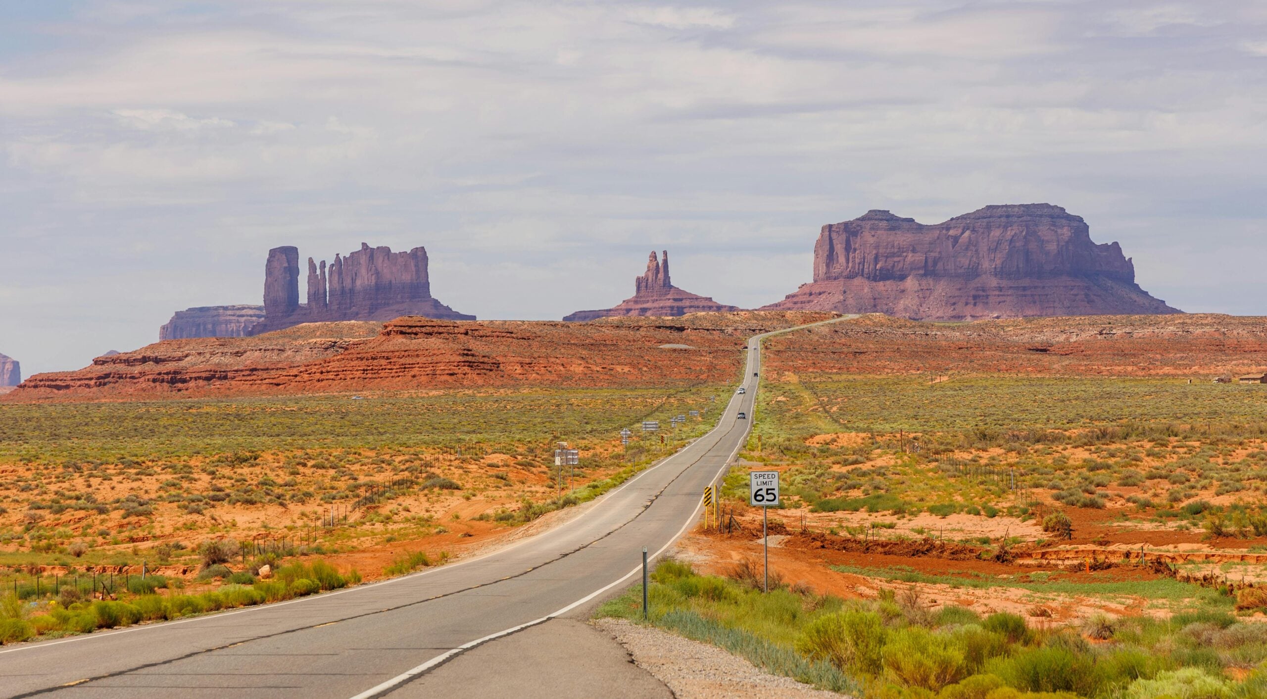 A long, straight road stretches into the desert toward towering red rock formations under a partly cloudy sky, with a speed limit sign reading 65 mph along the roadside.