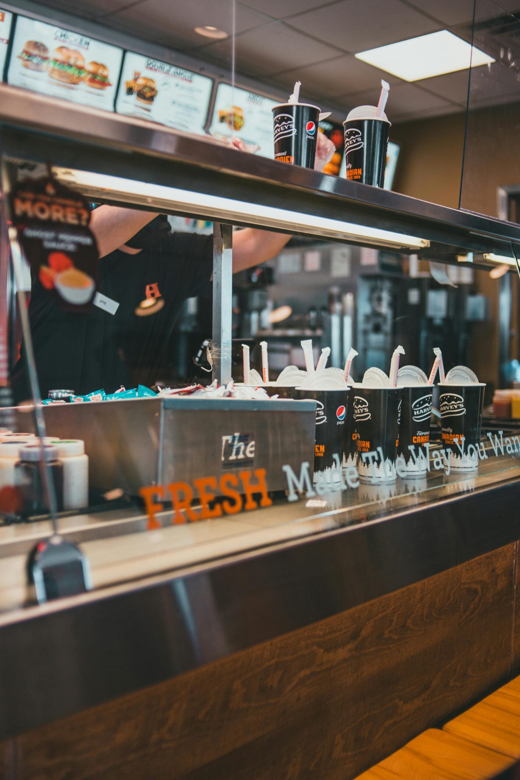 A fast food worker prepares drinks behind a counter, with several large cola cups and lids lined up. The menu boards and dining area are visible in the background through the glass.