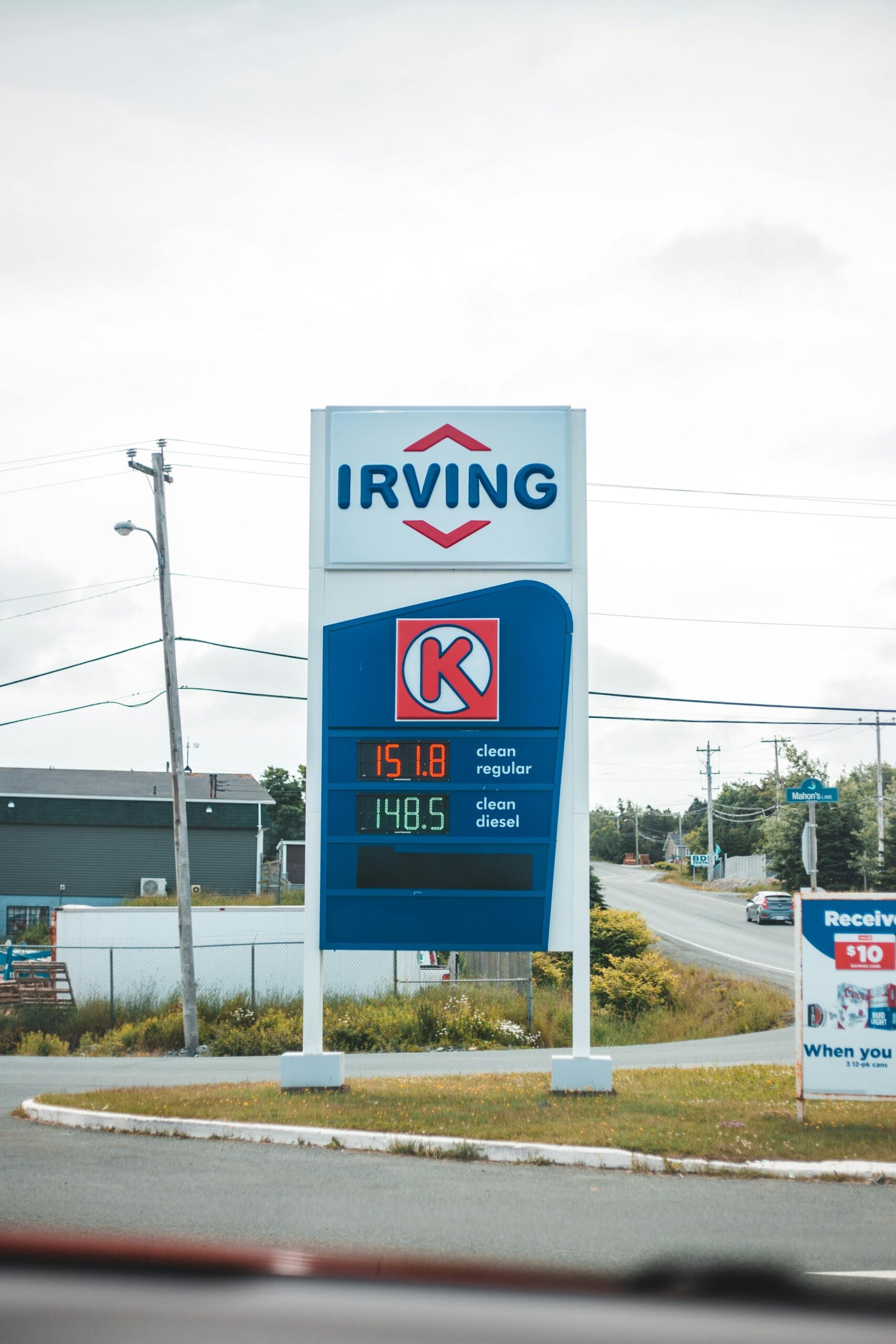 A roadside Irving gas station sign displays fuel prices: regular at 151.8 and diesel at 148.5. Power lines, buildings, and a partly cloudy sky are visible in the background.