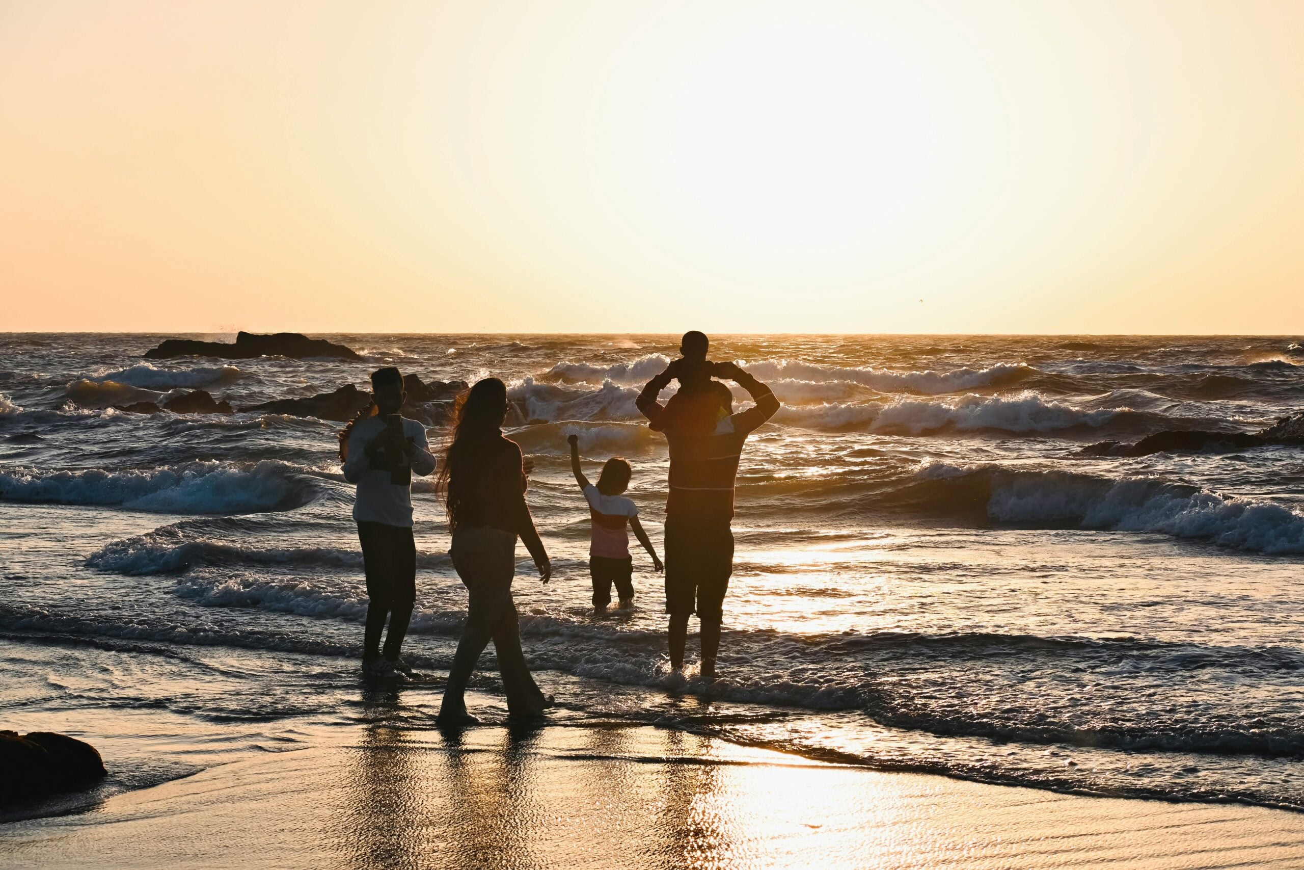 A family walks along the shoreline at sunset, with waves crashing in the background. One adult carries a child on their shoulders while others walk nearby, all silhouetted against the glowing sky and reflecting water.