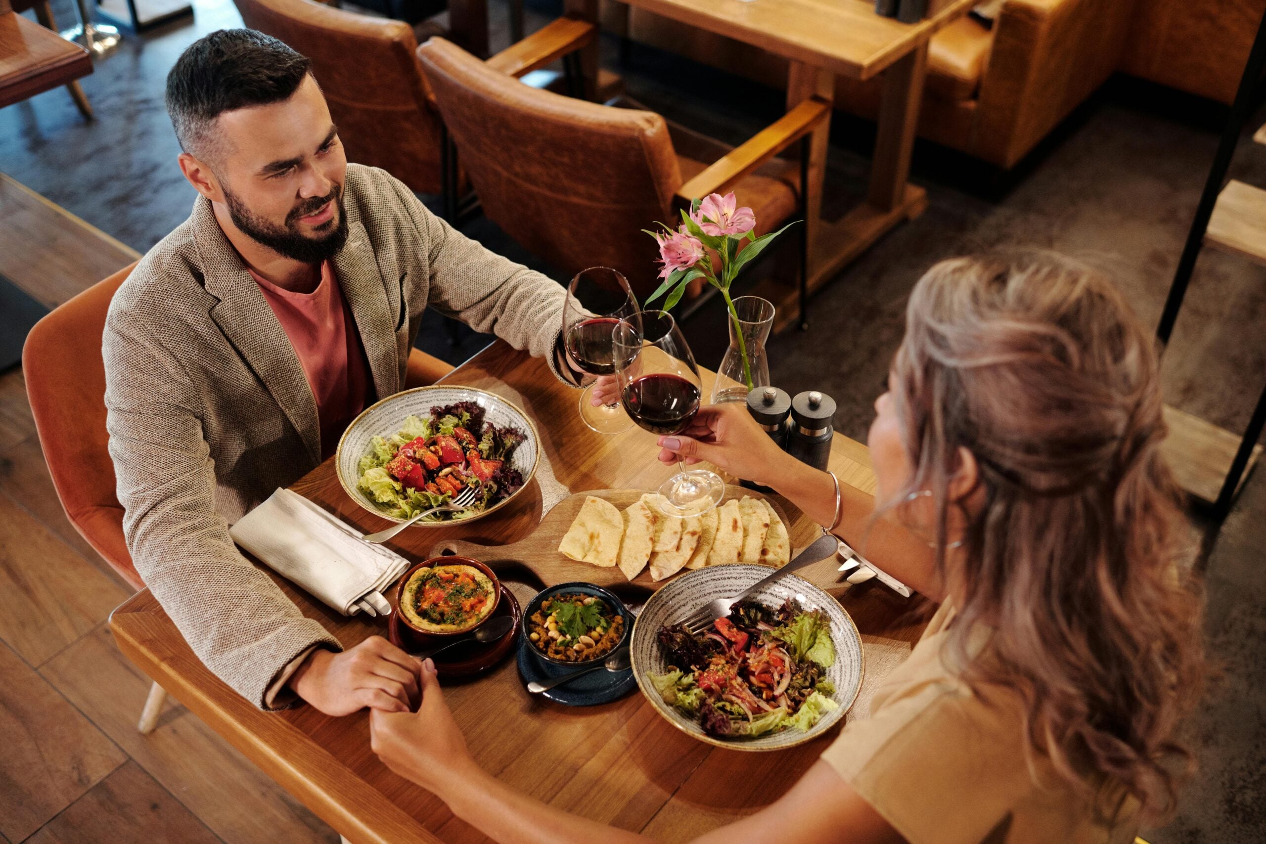 A man and woman sit at a restaurant table, holding hands and clinking glasses of red wine. The table is set with salads, bread, and appetizers. The atmosphere is warm and inviting, with a pink flower in a vase.