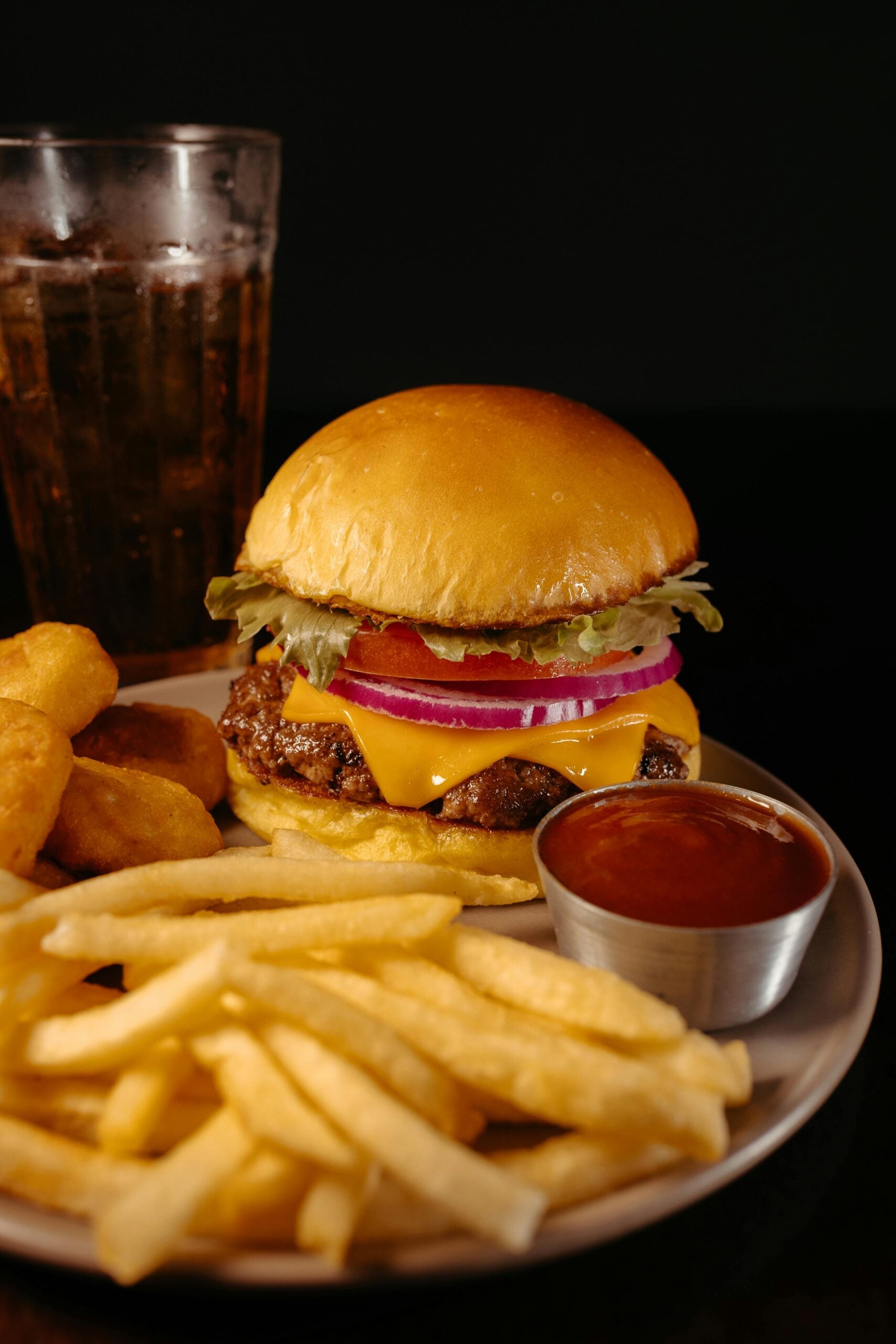 A plate with a cheeseburger topped with lettuce, tomato, onion, and cheese, served with French fries, nuggets, a cup of dipping sauce, and a glass of soda against a dark background.