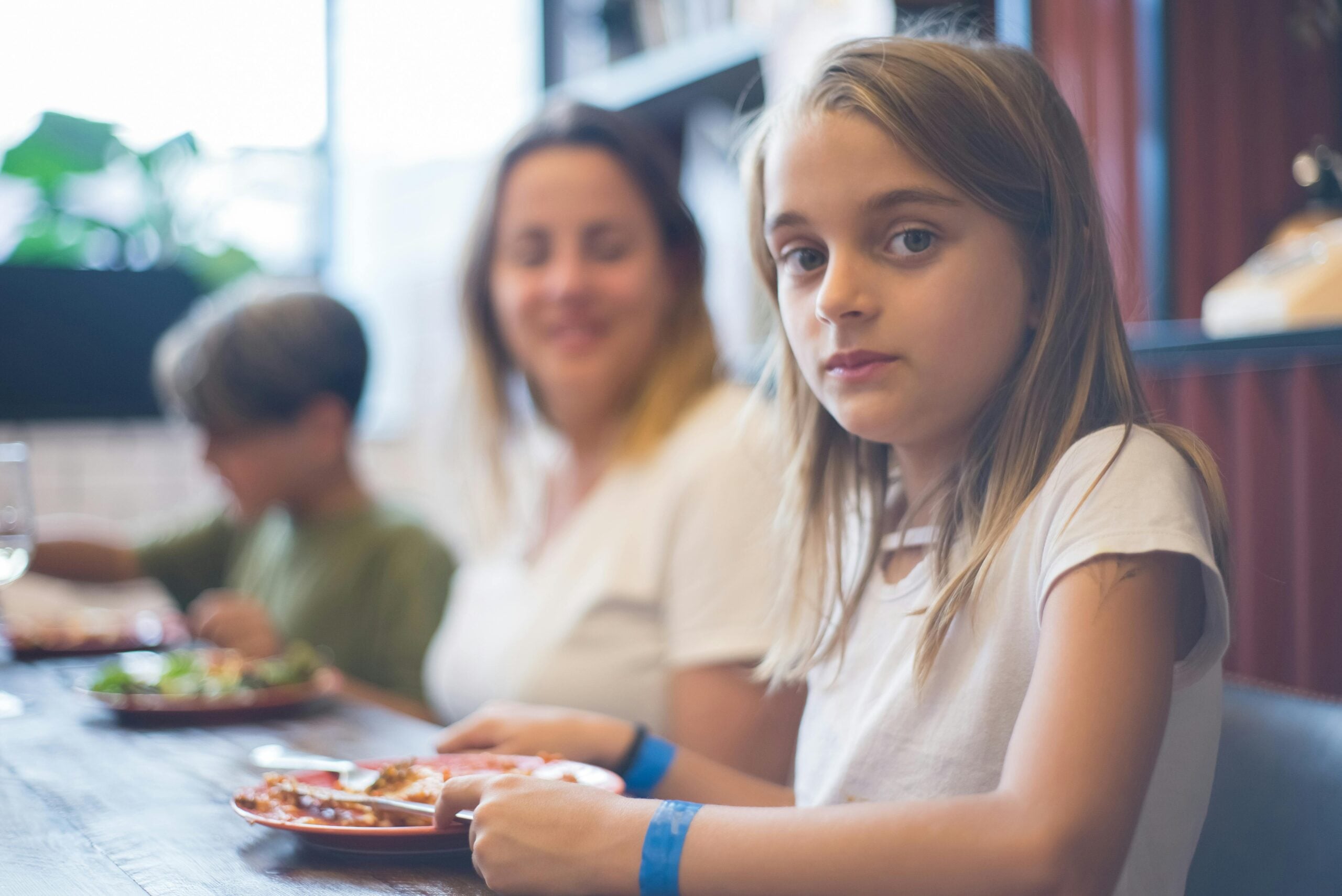 A young girl with long blonde hair sits at a table holding a plate of food, looking at the camera. In the background, a woman and another child are eating, with soft natural light coming from a window.