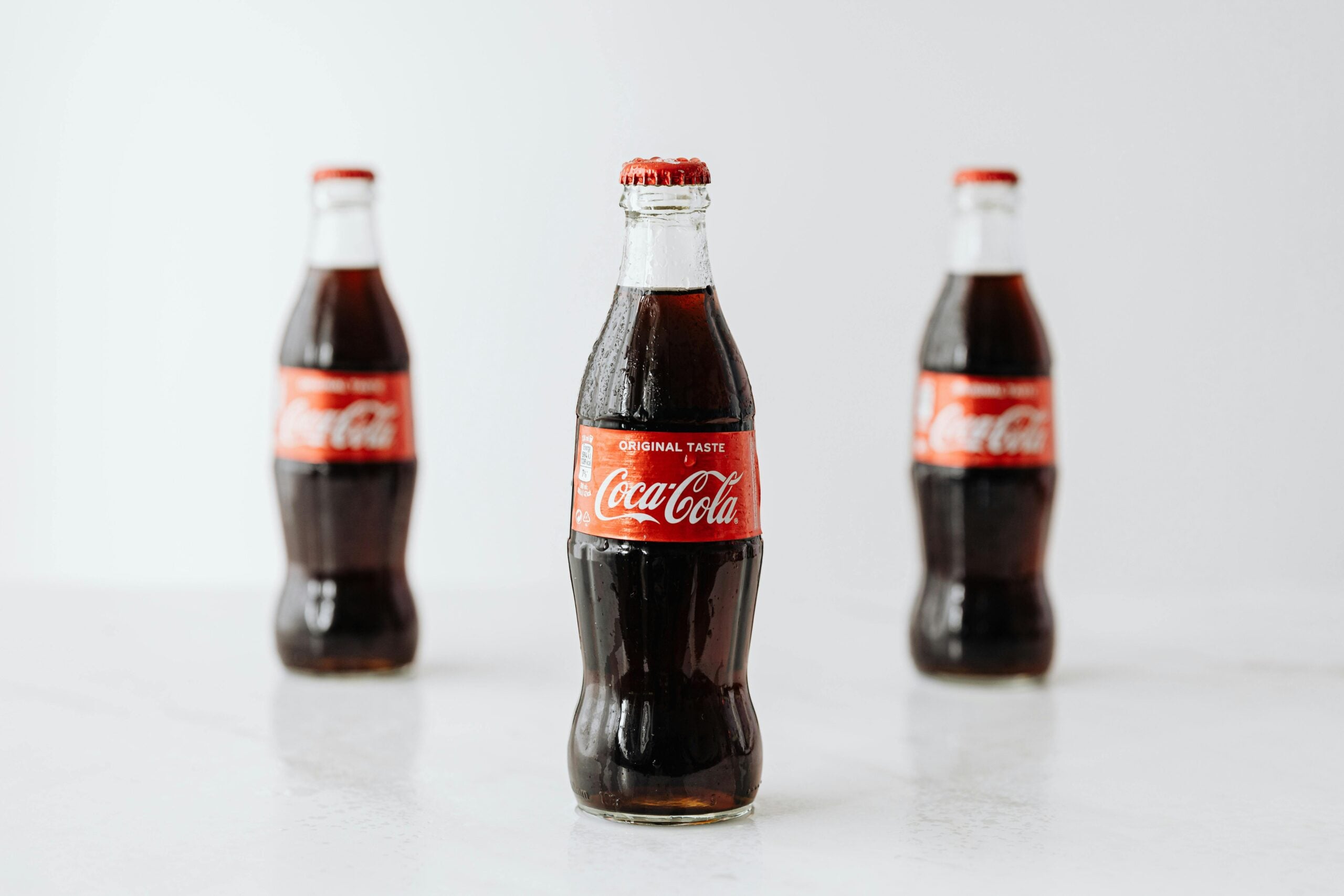 Three classic glass Coca-Cola bottles with red labels are placed on a white surface. The bottle in the center is in focus, while the two bottles in the background are slightly blurred.