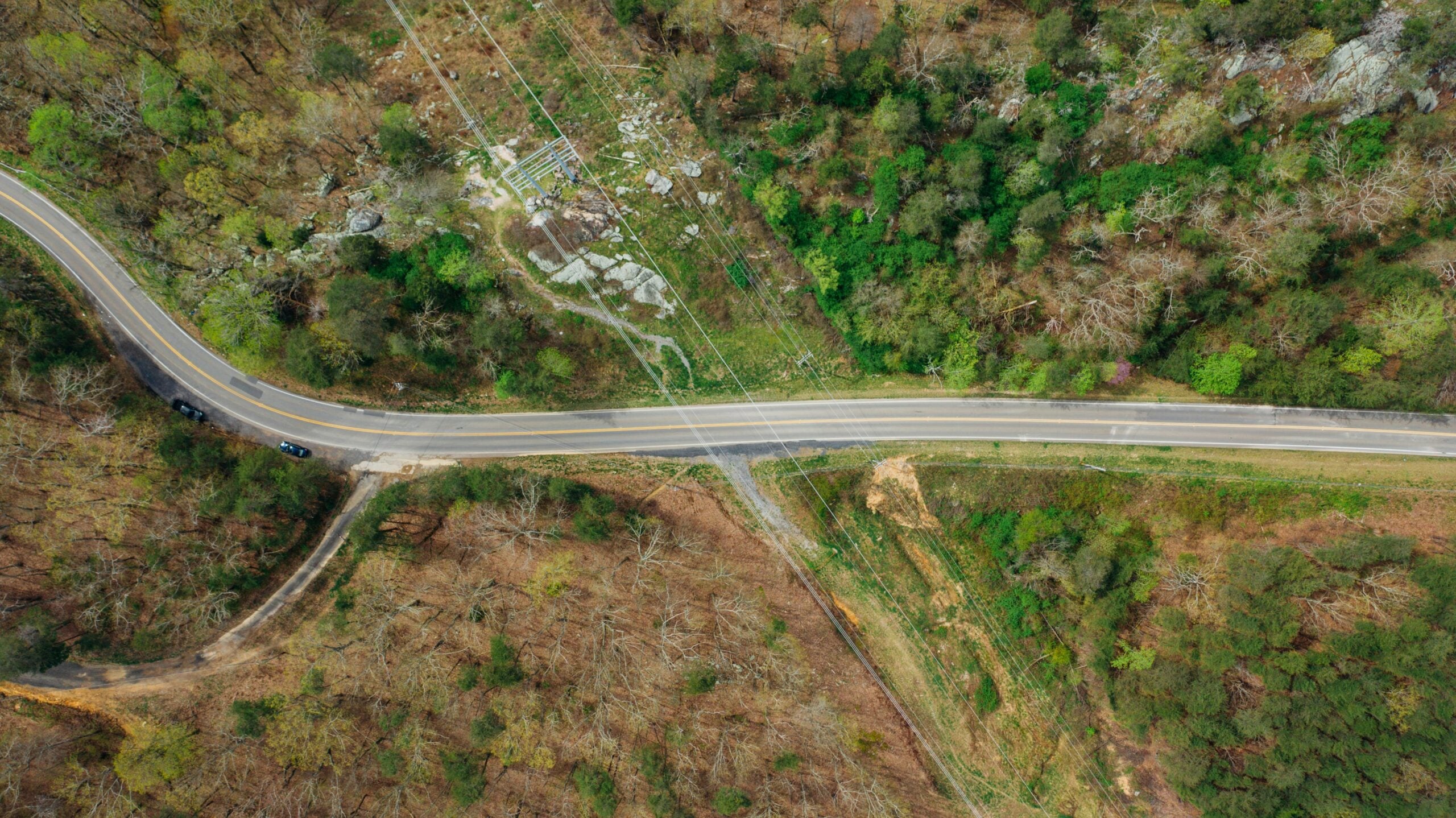 Aerial view of a curved road cutting through a forested area, with patches of green and brown trees on either side and small rocky clearings visible near the road.