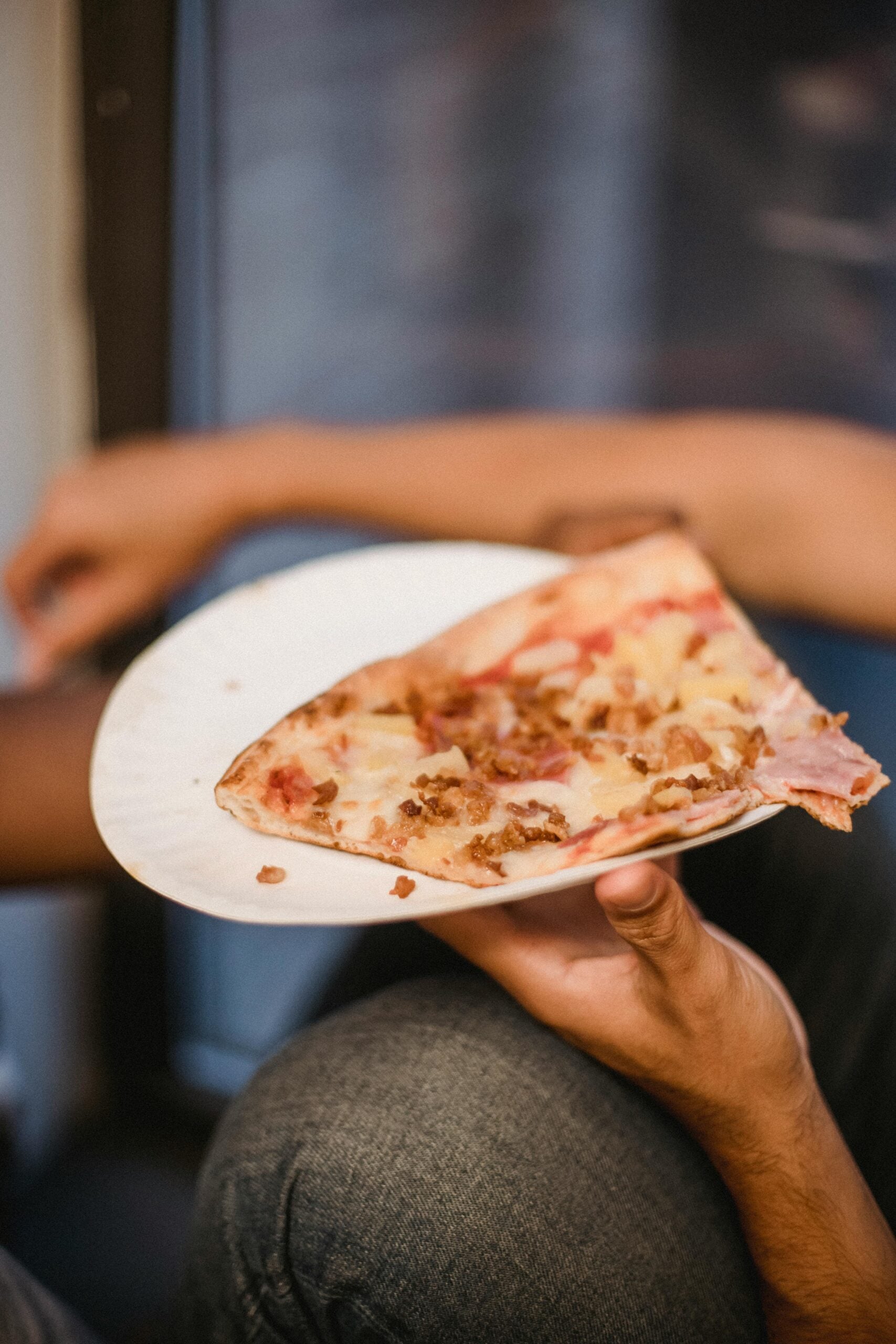 A person holds a white paper plate with two large slices of pizza topped with cheese and bits of meat, resting on their lap.
