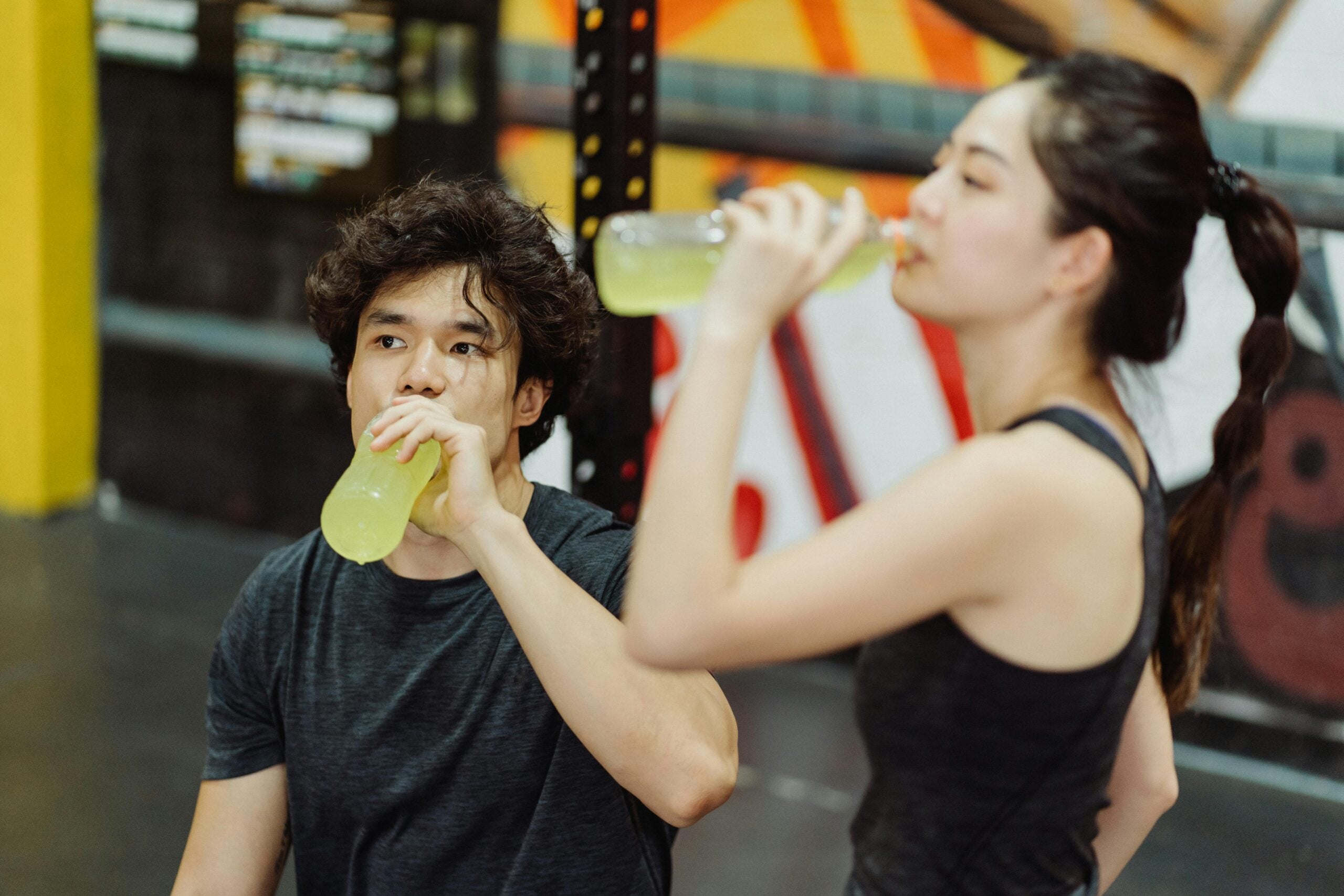 Two people in workout clothes drink yellow sports drinks in a gym. One person sits and looks ahead while the other stands, both appearing to take a break from exercising. Gym equipment and colorful wall art are visible in the background.