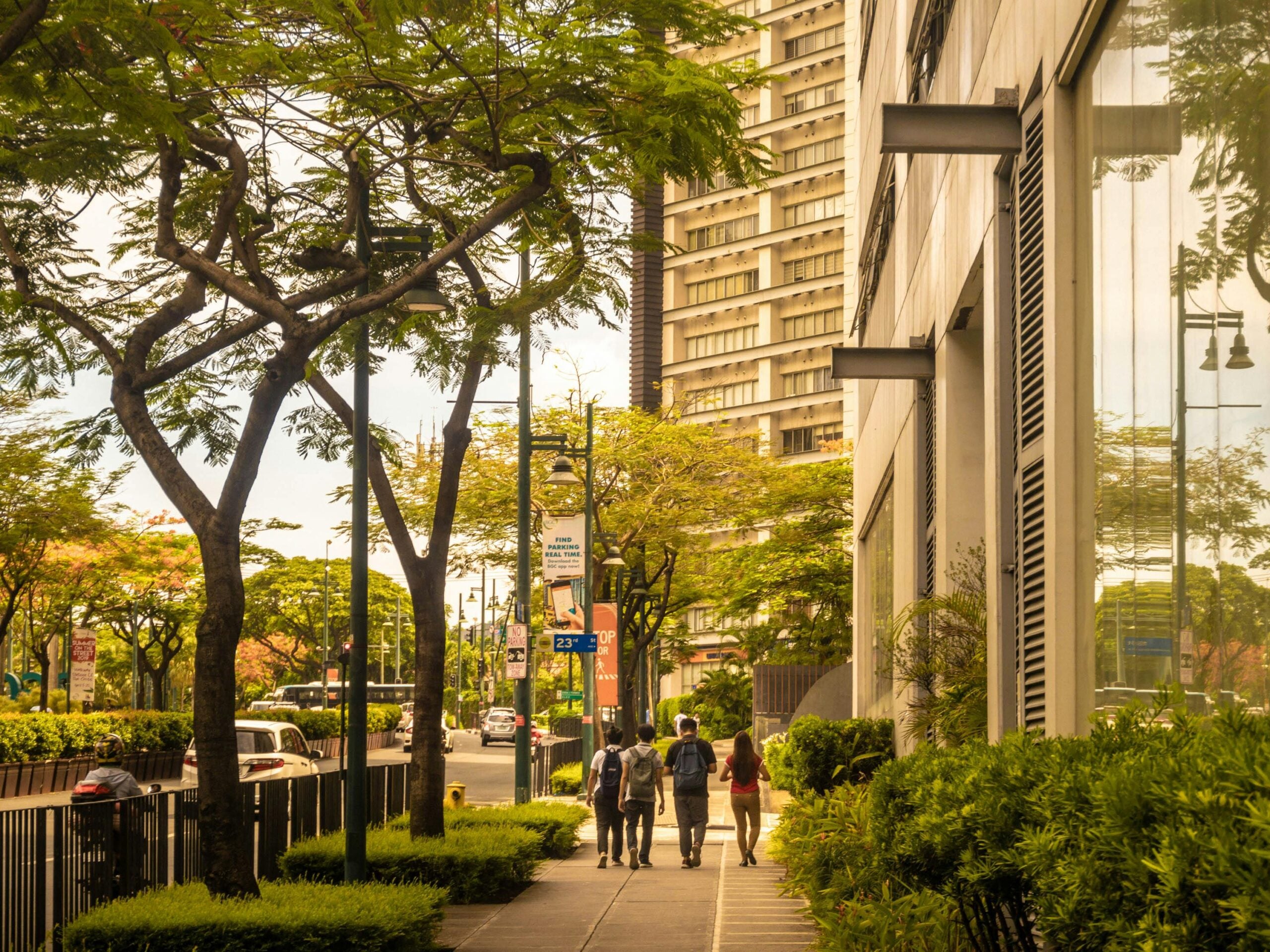 Cheap Airbnb and hotel tricks. People walk along a tree-lined city sidewalk next to a modern building with large windows. Tall buildings and greenery line the street under a warm, sunny sky.