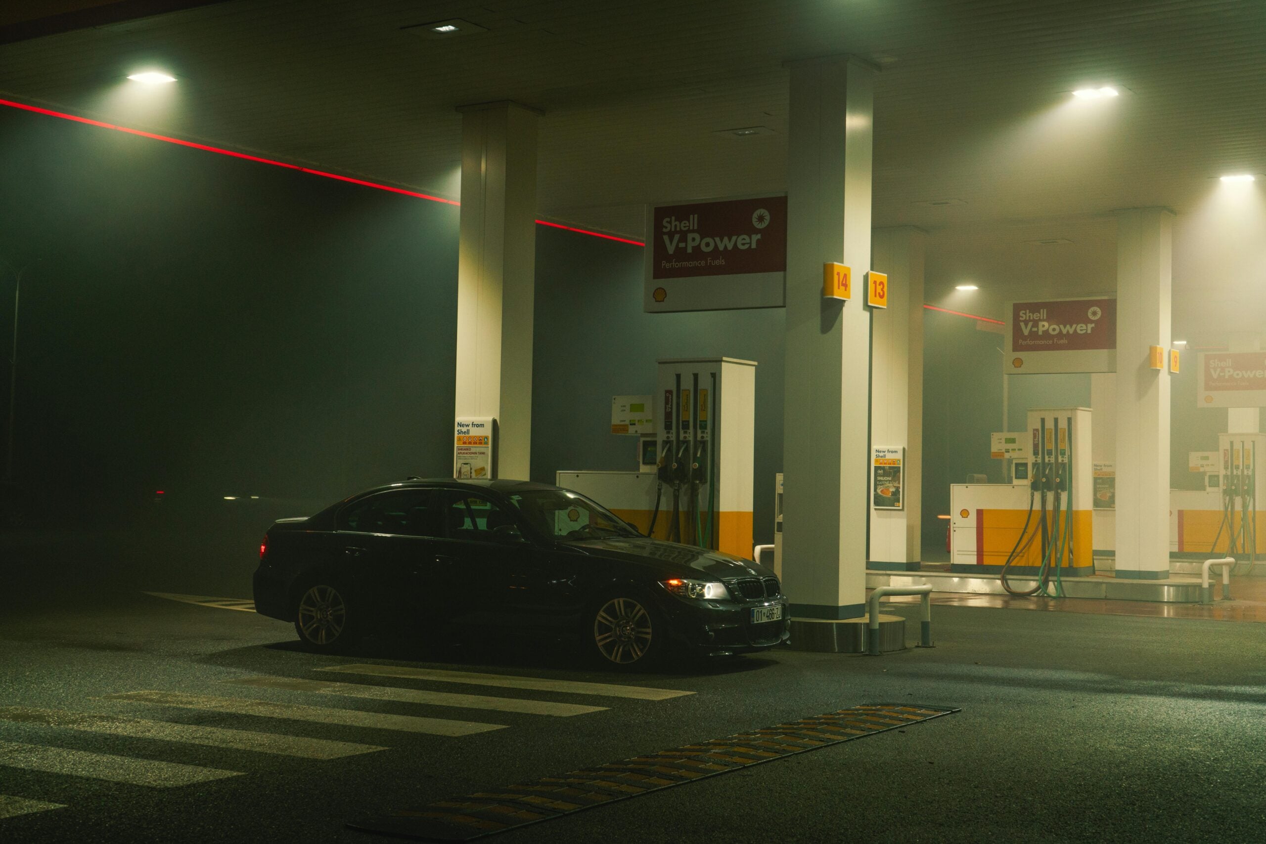 A black sedan is parked at a Shell gas station at night, illuminated by overhead lights with a misty atmosphere and empty surroundings. Multiple fuel pumps are visible under the station canopy.