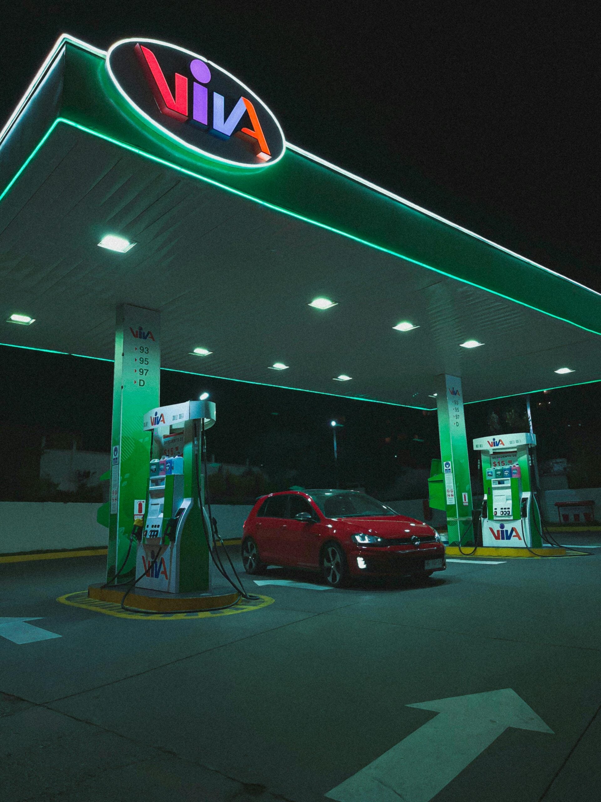 A red car is parked at a brightly lit VIVA gas station at night, under a large canopy with glowing green accents and fuel price signs visible on the pumps.