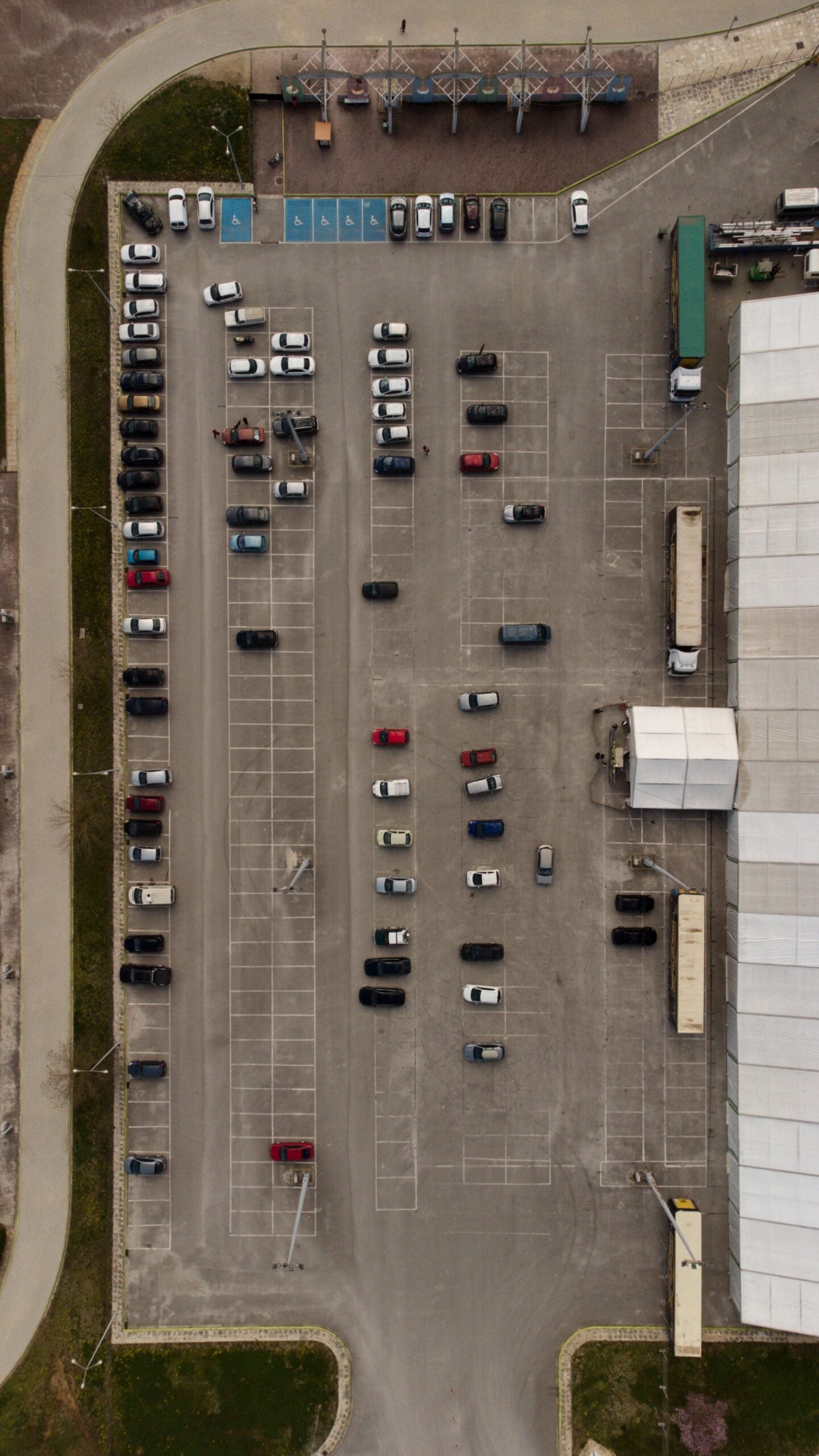 Aerial view of a parking lot with several rows of parked cars, some empty spaces, trucks near a large white industrial building, and handicap parking spots at the top left. Road curves along the parking lot’s edge.