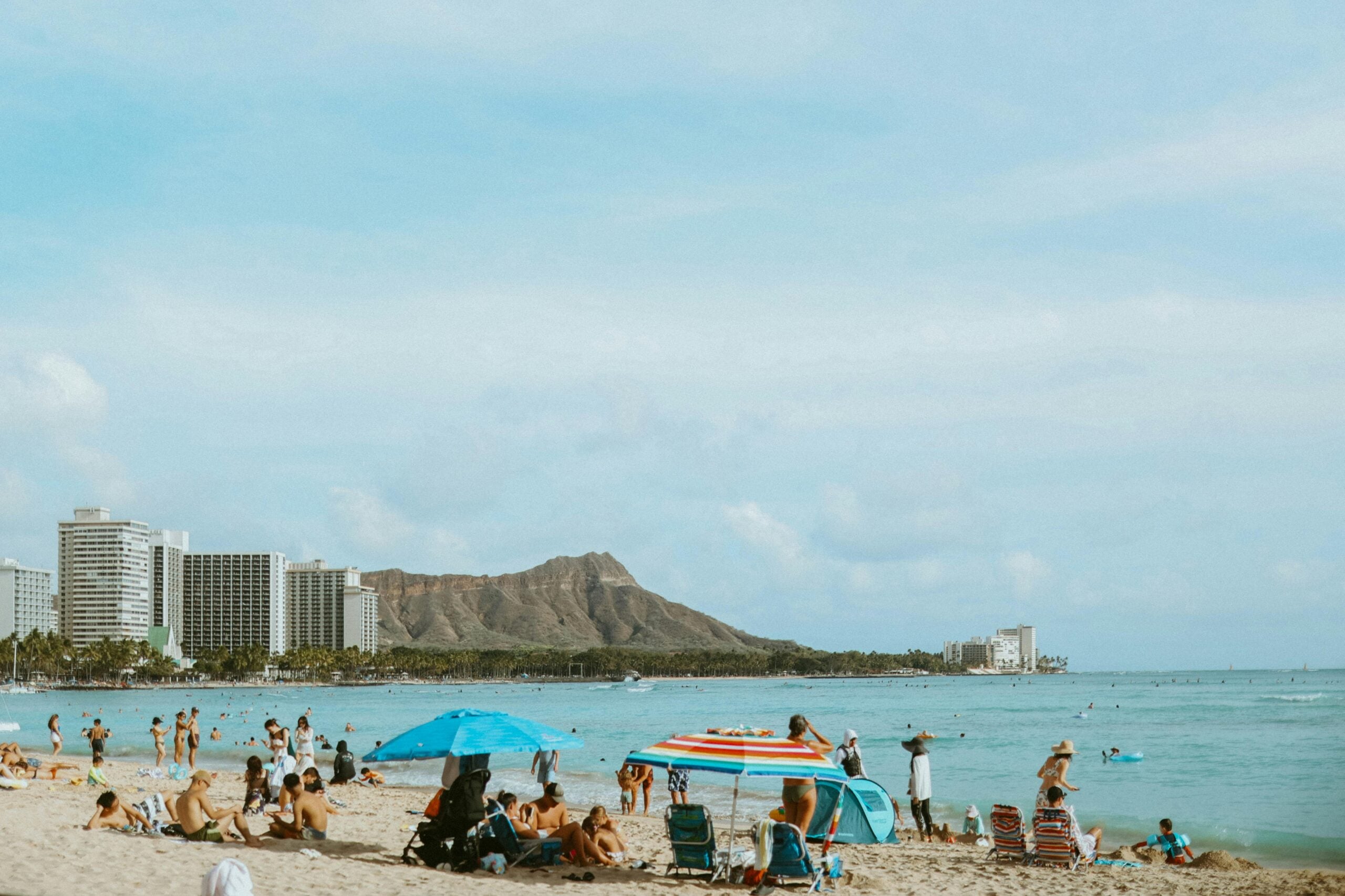 People relax on a sandy beach under umbrellas, with swimmers in the blue ocean. High-rise buildings and a green volcanic mountain are visible in the background under a partly cloudy sky.