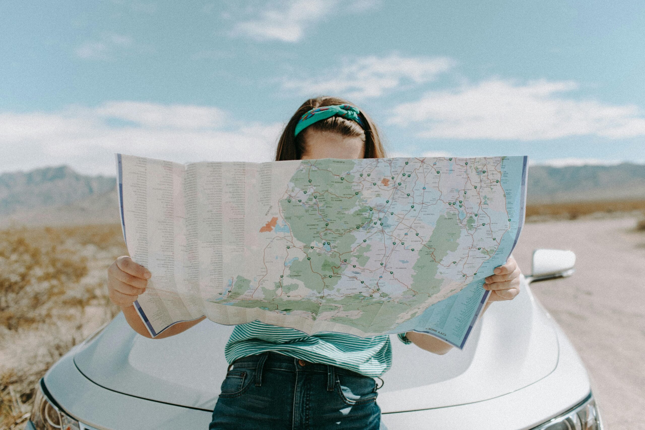A person sits on the hood of a white car in a desert landscape, holding and reading a large map that covers most of their face. Mountains and blue sky are visible in the background.