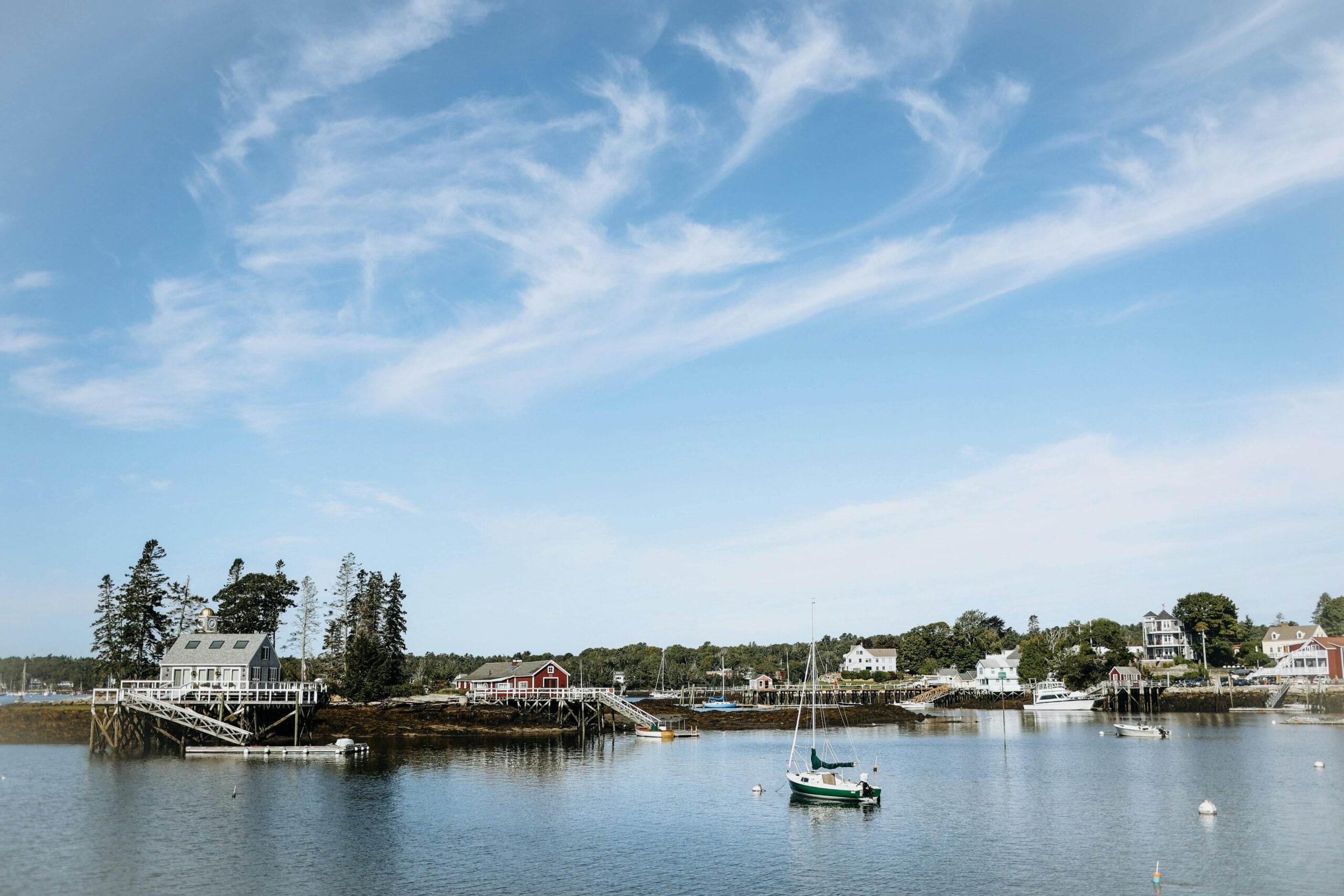 A scenic coastal village with houses, docks, and trees along the shoreline, sailboats and small boats floating on calm water, and wispy clouds in a blue sky.