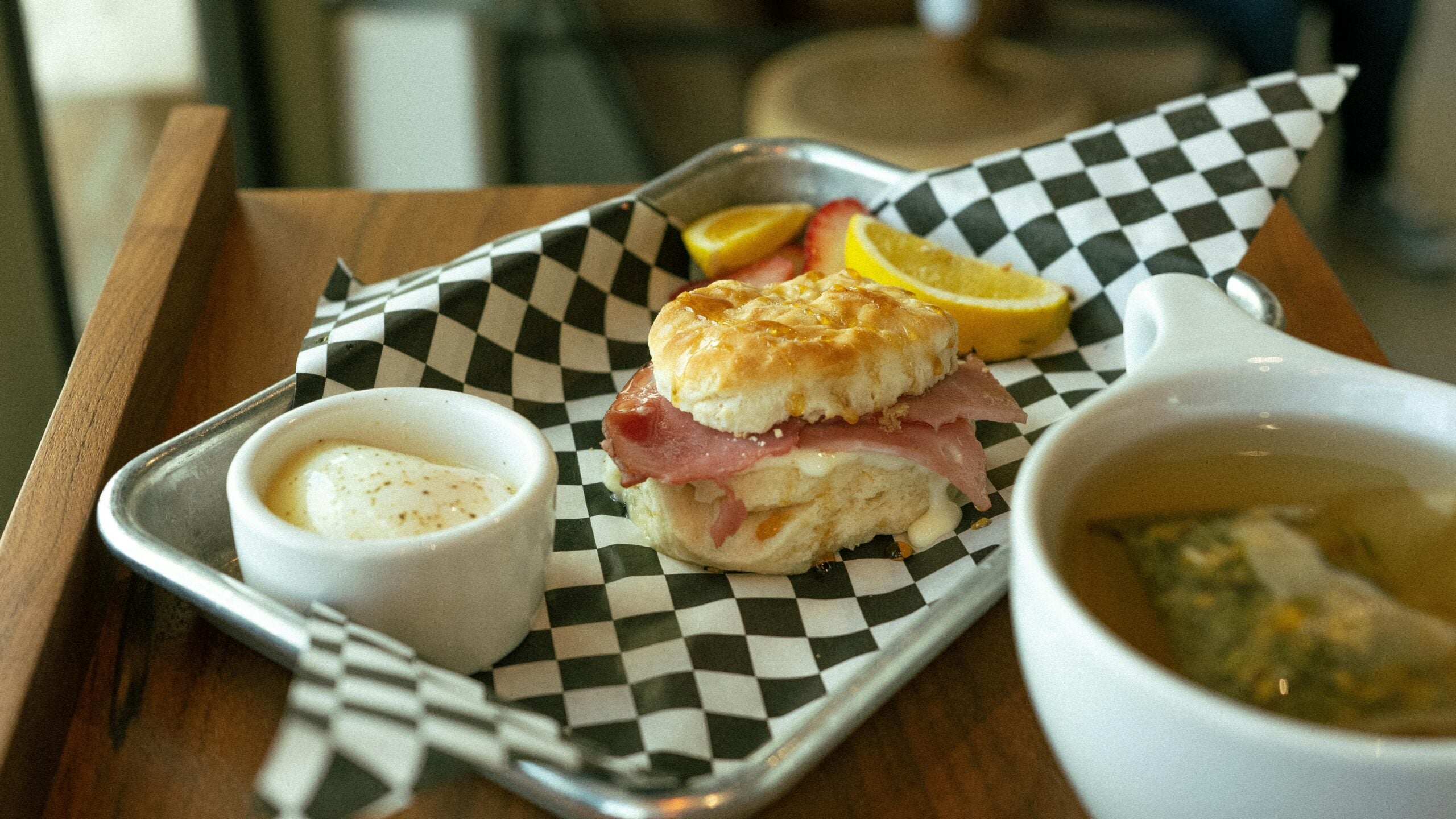 A breakfast sandwich with ham and egg on a biscuit sits on a checkered paper-lined tray, accompanied by lemon slices, a small cup of butter, and a mug of tea.