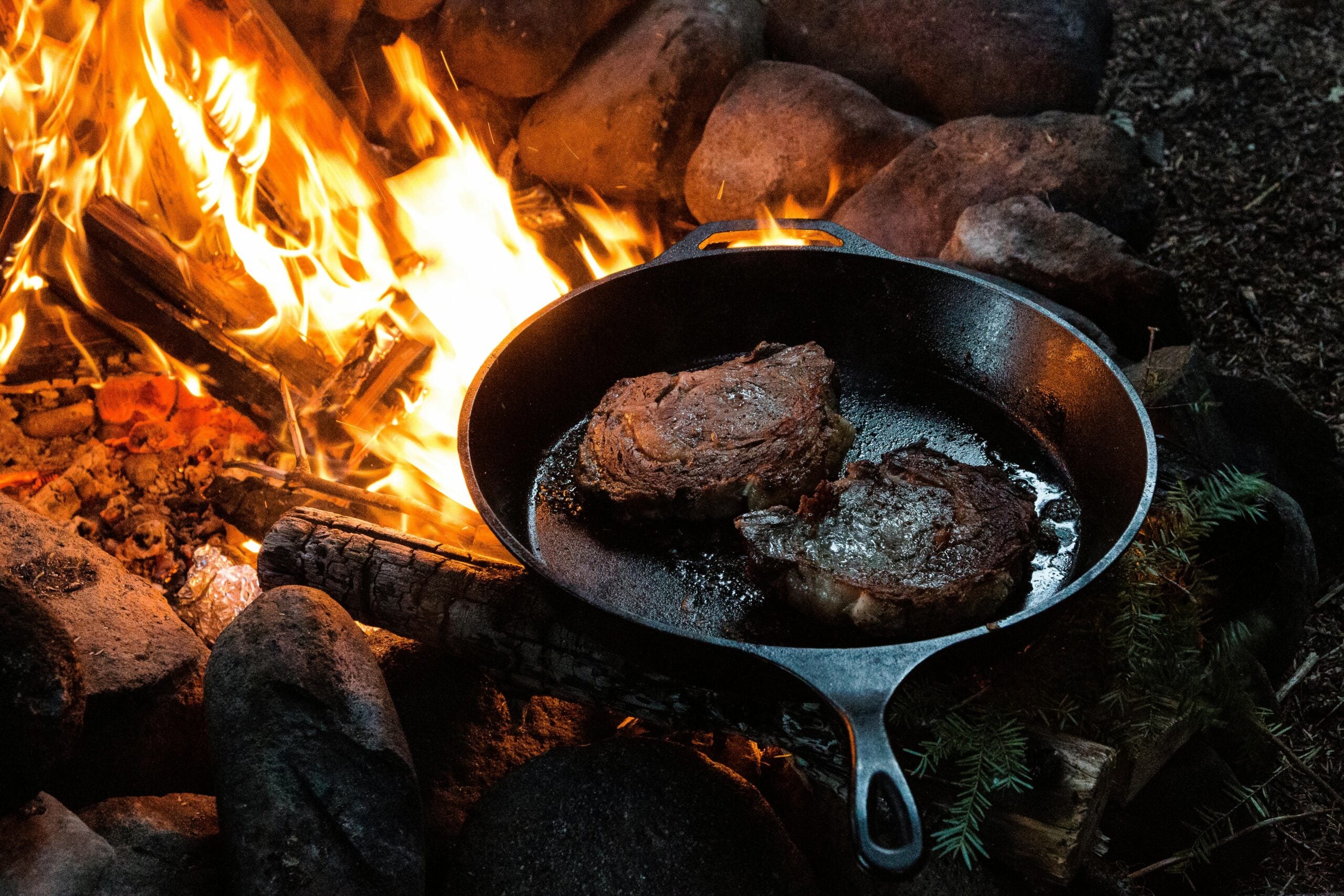 Two steaks cooking in a cast iron skillet over an open campfire, surrounded by rocks and burning logs, creating a rustic outdoor cooking scene.