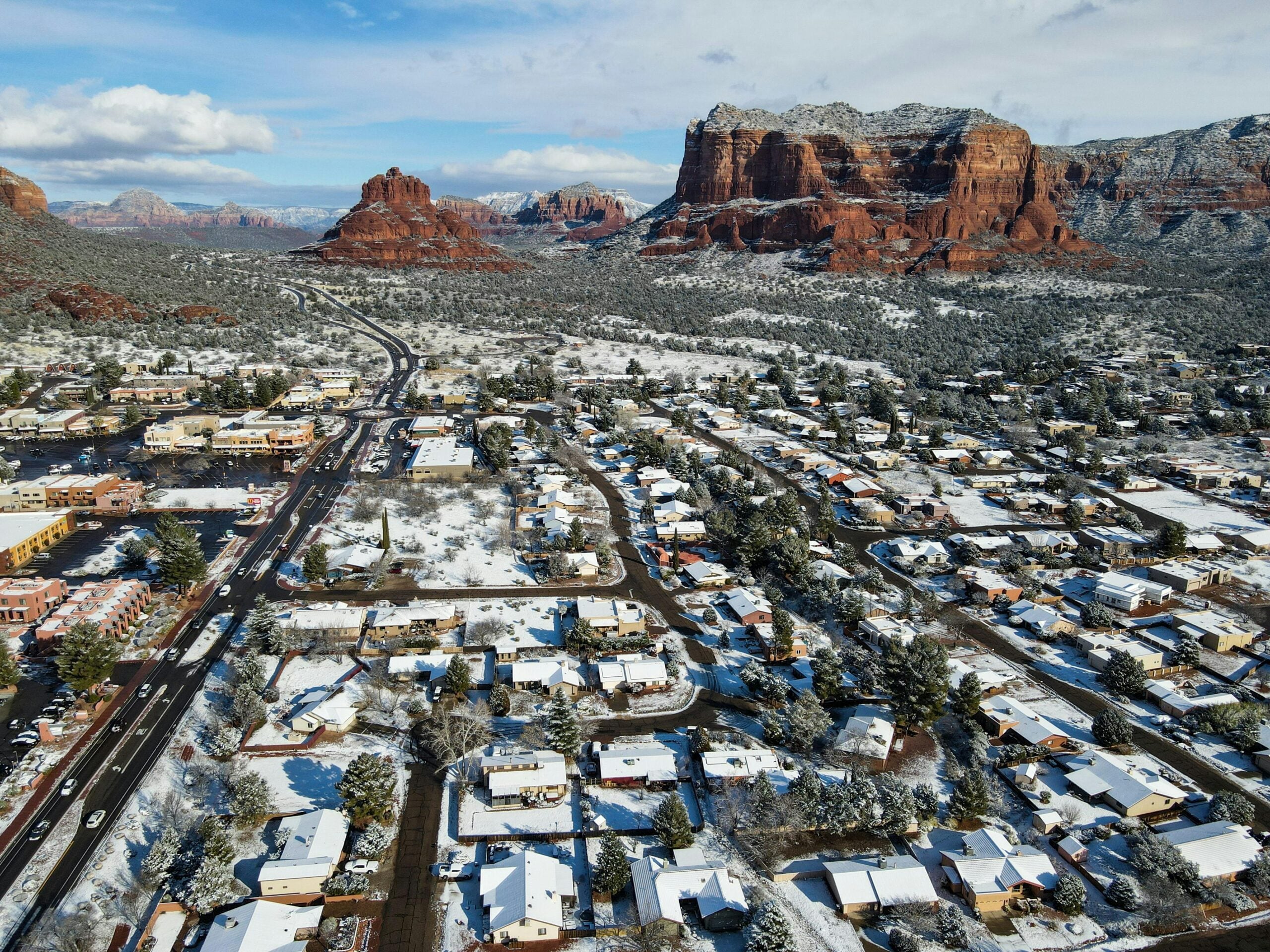 Aerial view of a suburban neighborhood covered in a light layer of snow with red rock formations and mesas in the background under a partly cloudy sky.