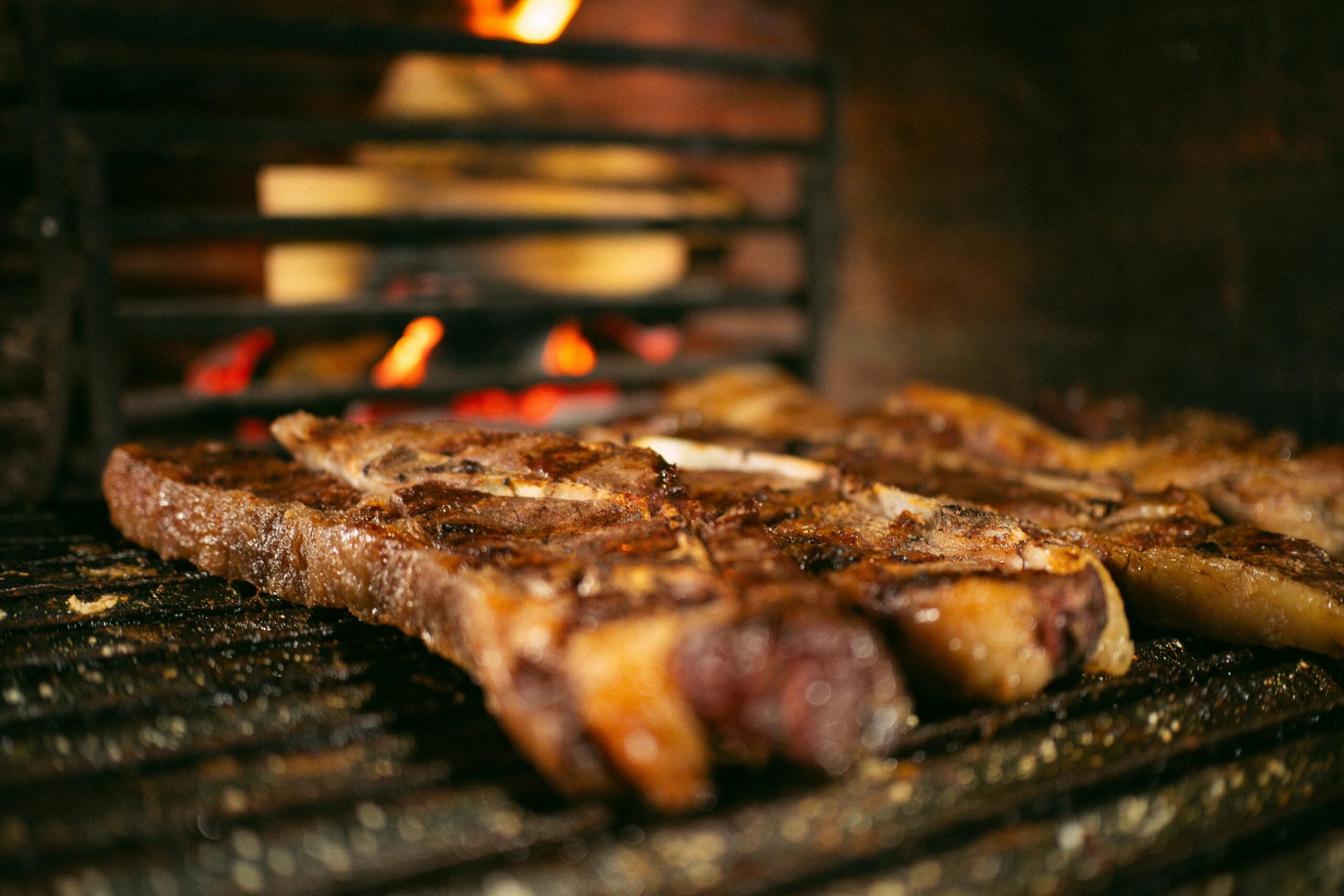 Close-up of juicy, grilled steaks cooking on a barbecue grill with flames and glowing coals in the background, highlighting the charred surface and sizzling fat.