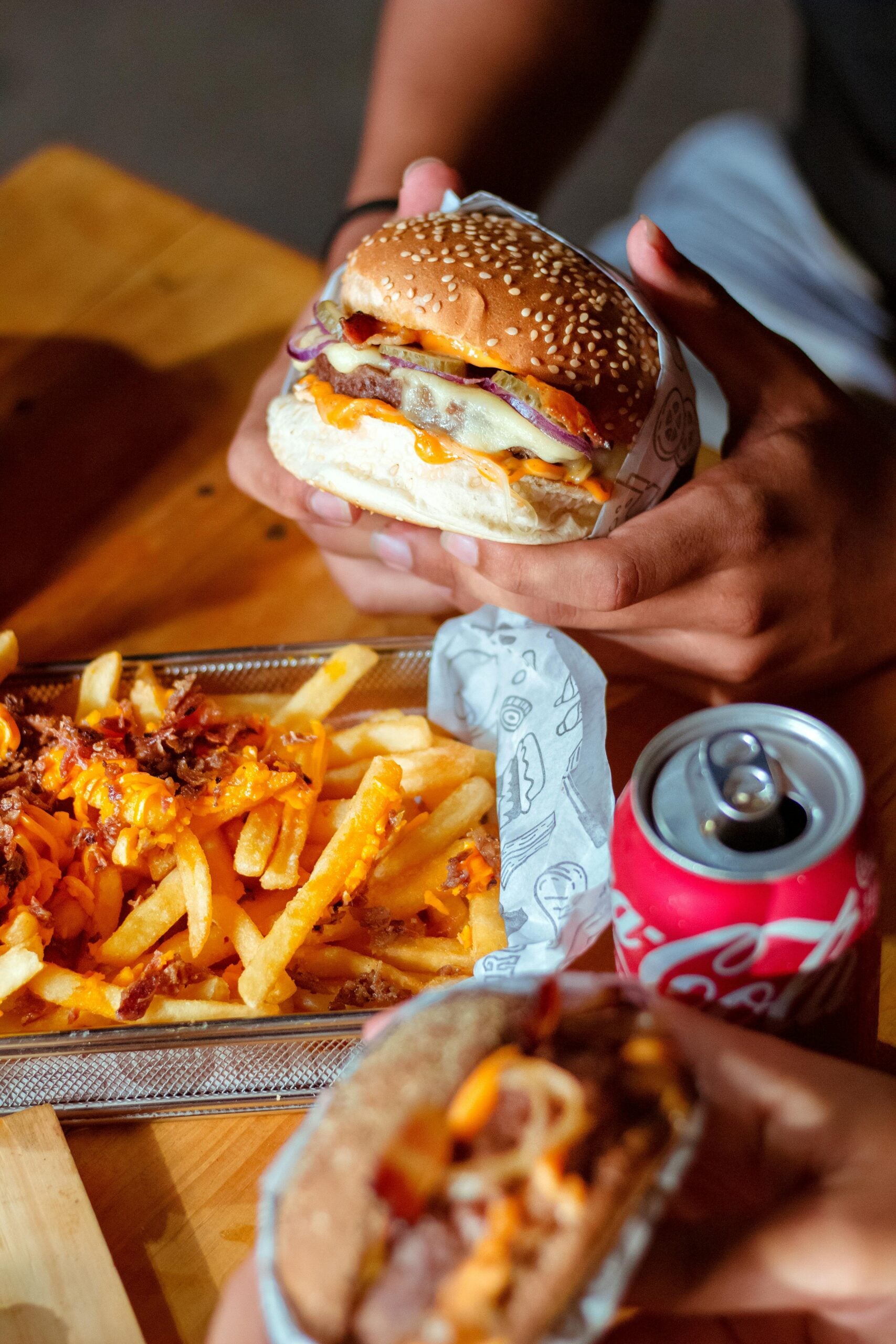 Two hands holding cheeseburgers with bacon and sauce over a wooden table, next to a basket of loaded fries topped with cheese and bacon, and an open can of Coca-Cola.