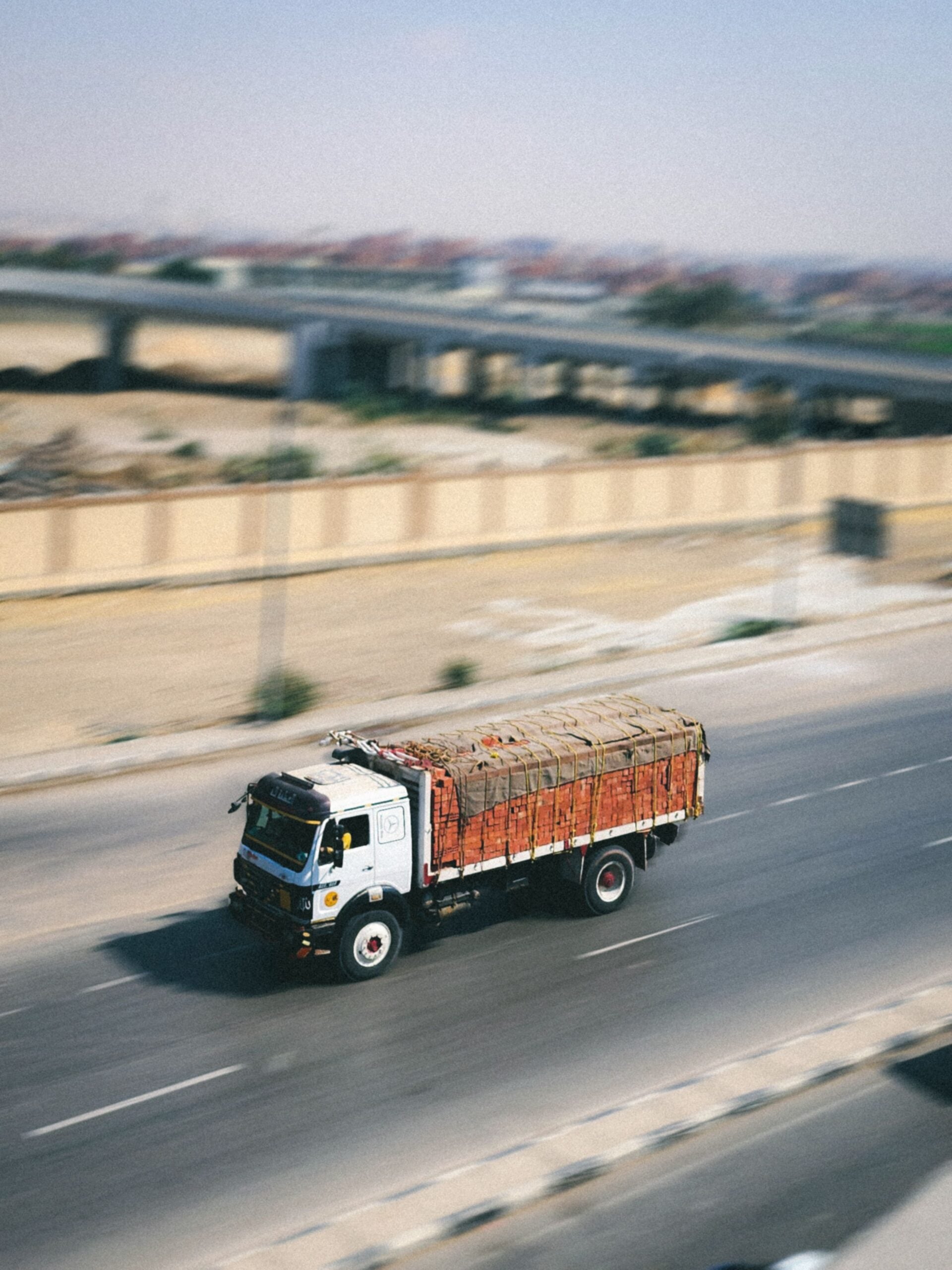 A white and red truck loaded with goods drives along a wide, empty road with a fence and overpasses in the blurred background, suggesting motion and speed.