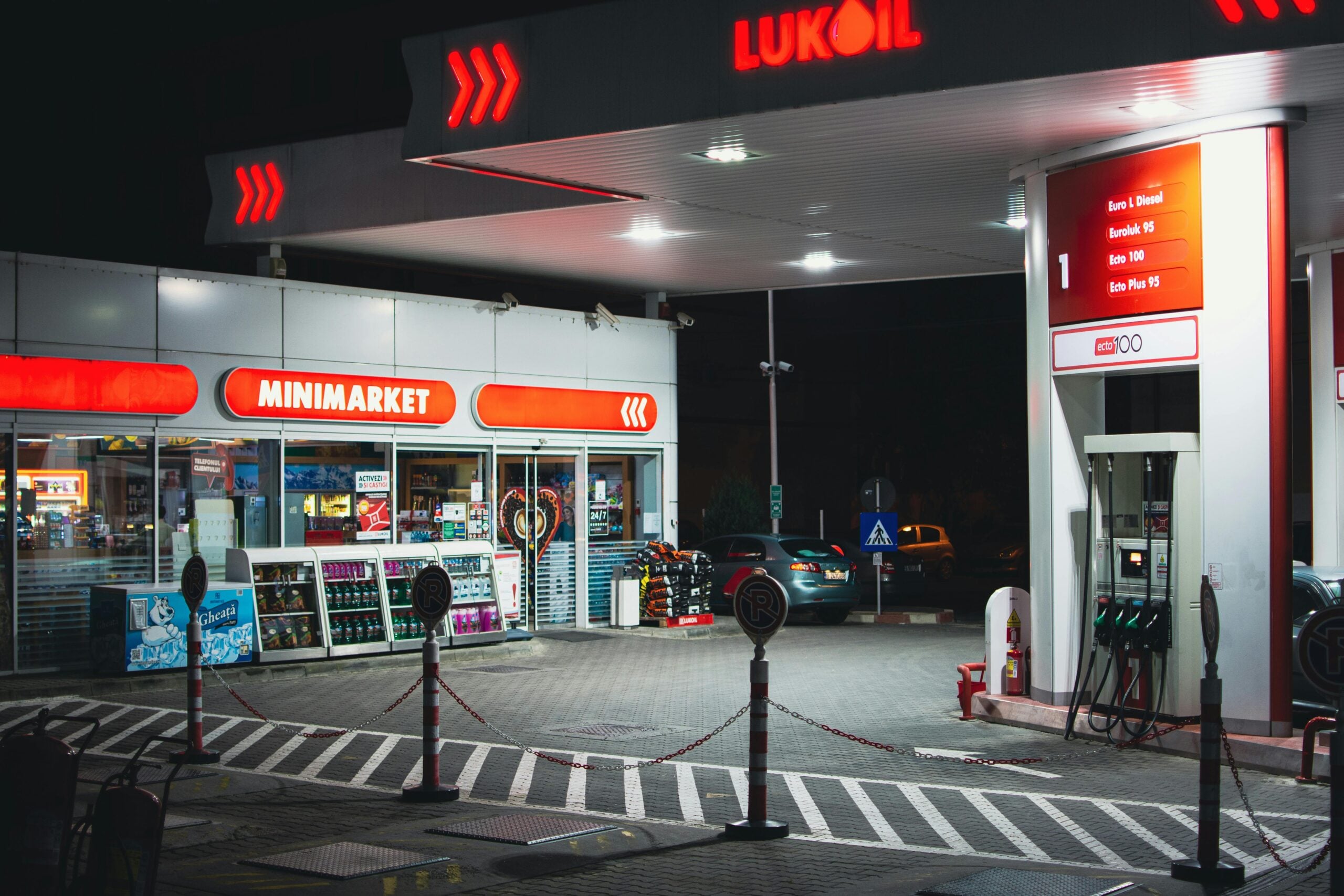 A Lukoil gas station at night with a minimarket, illuminated signs, fuel pumps, and a parked car. The area is clean and well-lit, with visible safety markings on the ground.