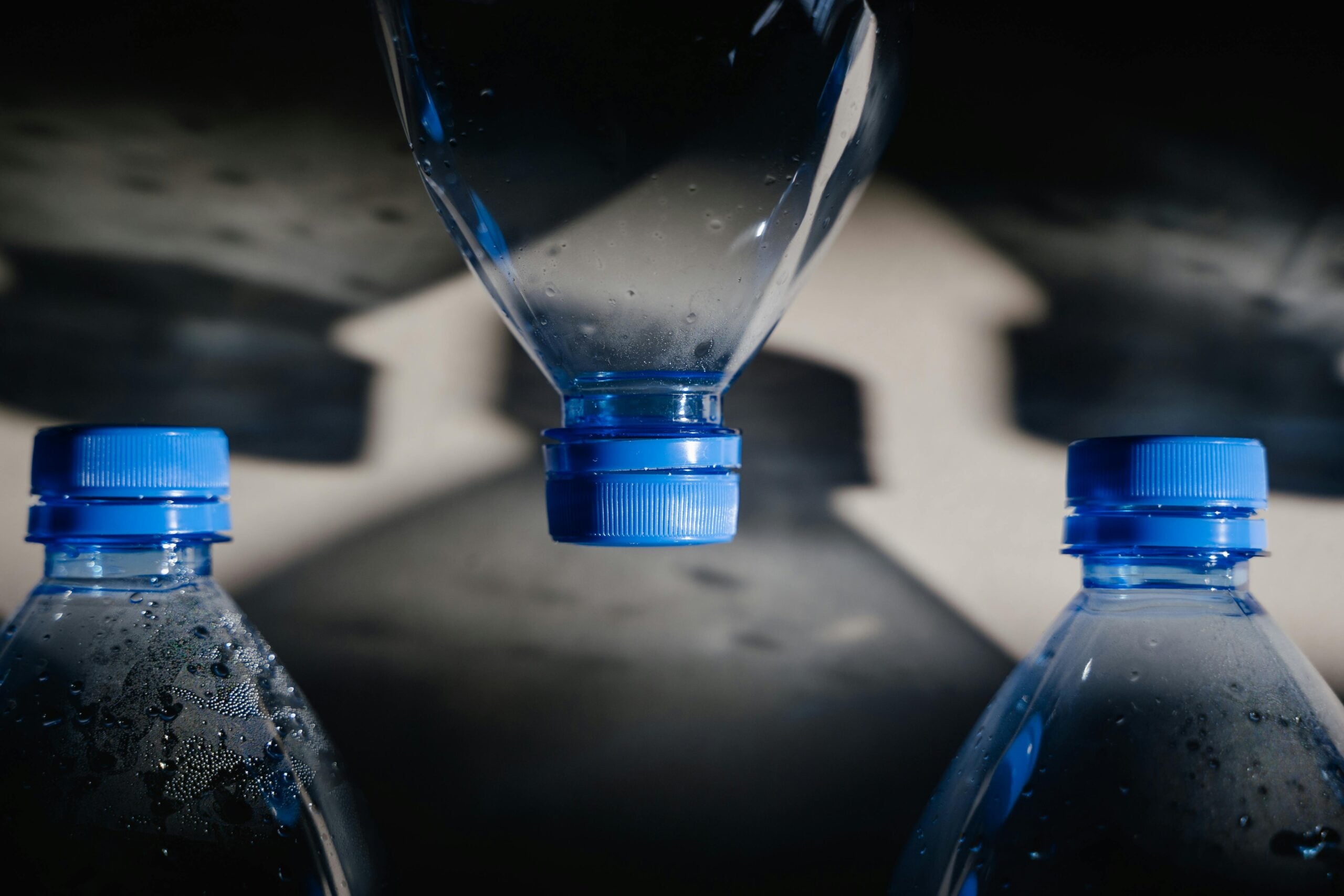 Close-up of three plastic water bottles with blue caps, two upright and one upside down in the center, with dramatic lighting creating distinct bottle-shaped shadows on a gray background.