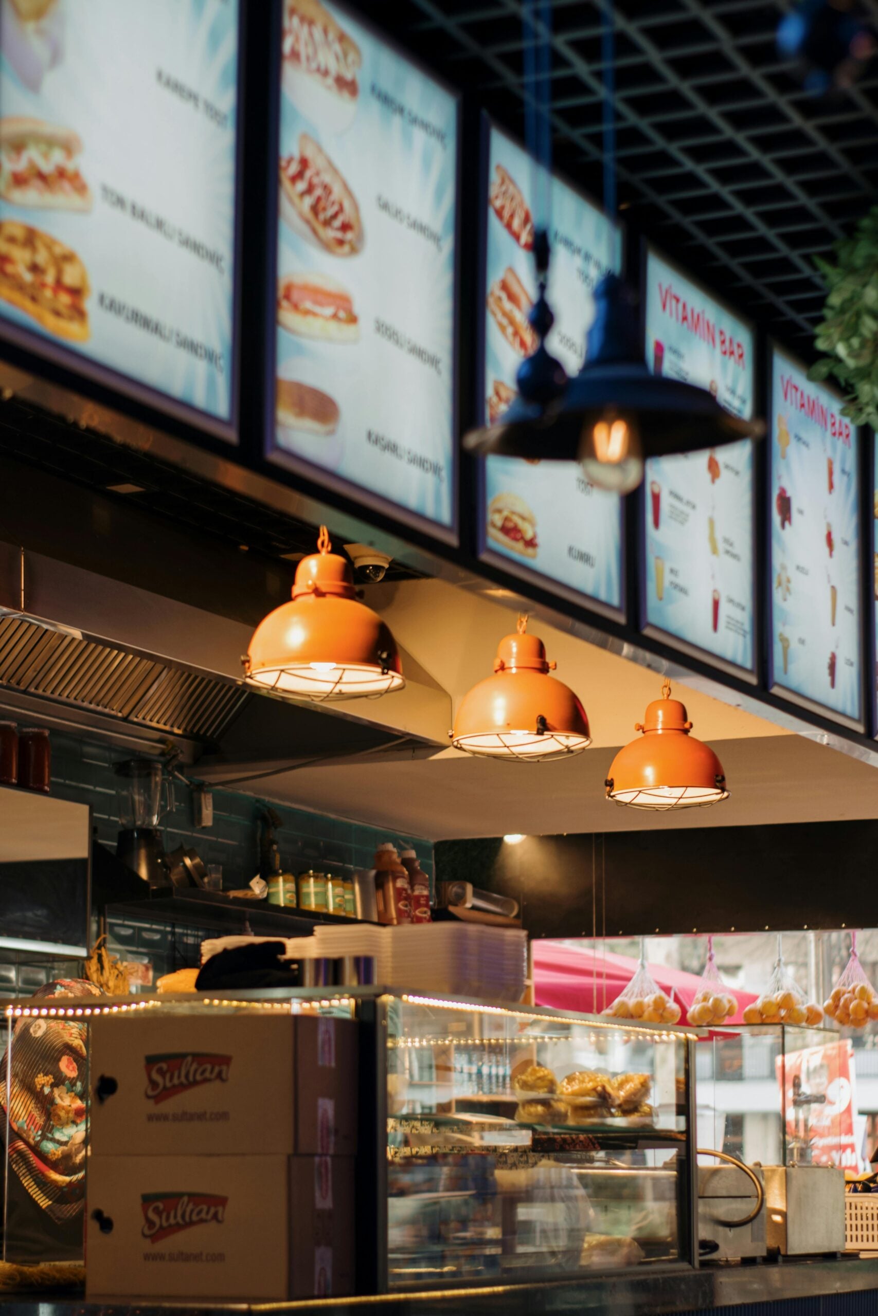 A brightly lit fast food restaurant counter with hanging orange lamps, menu boards displaying food options above, and stacked boxes labeled "Sultan" in the foreground.