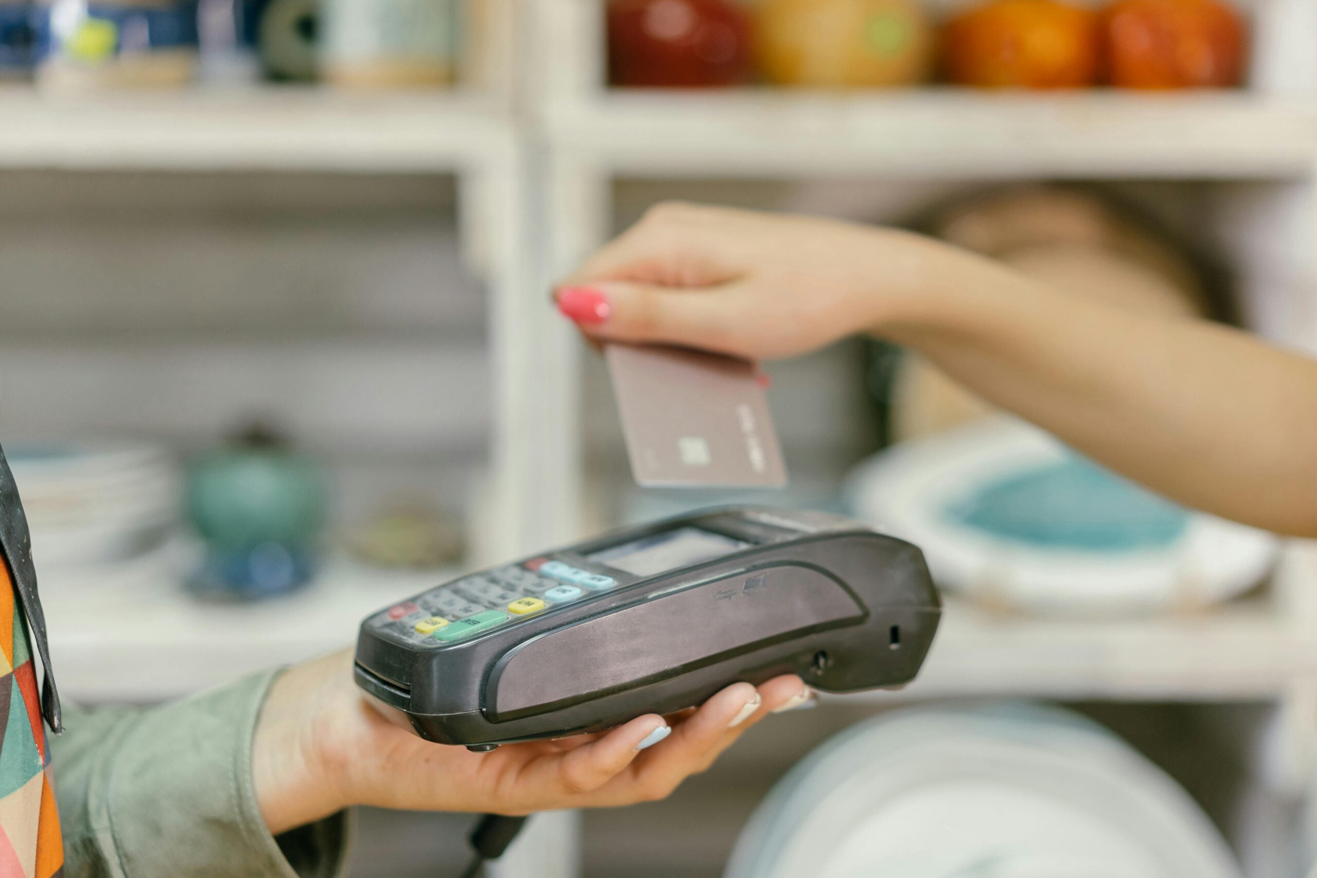 A person holds a payment terminal while another person pays by holding a credit card above it, demonstrating a contactless payment. Shelves with various items are blurred in the background.