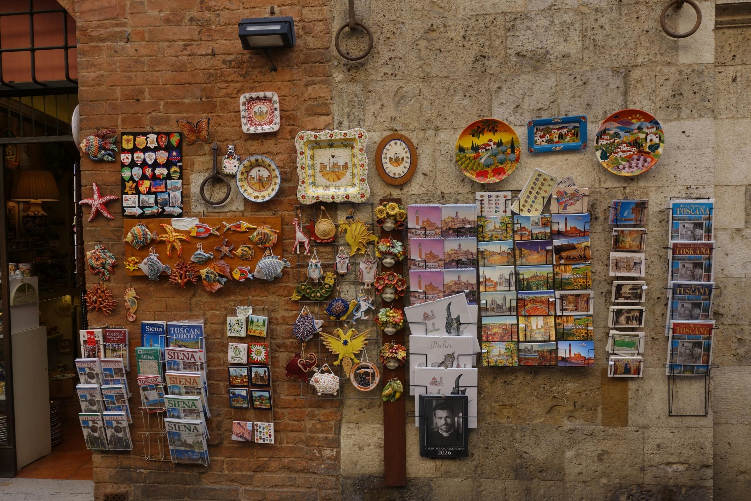 A shop wall displays colorful ceramic plates, magnets, decorative items, and racks of postcards featuring various scenes, likely from Siena and Tuscany, against a brick and stone exterior.