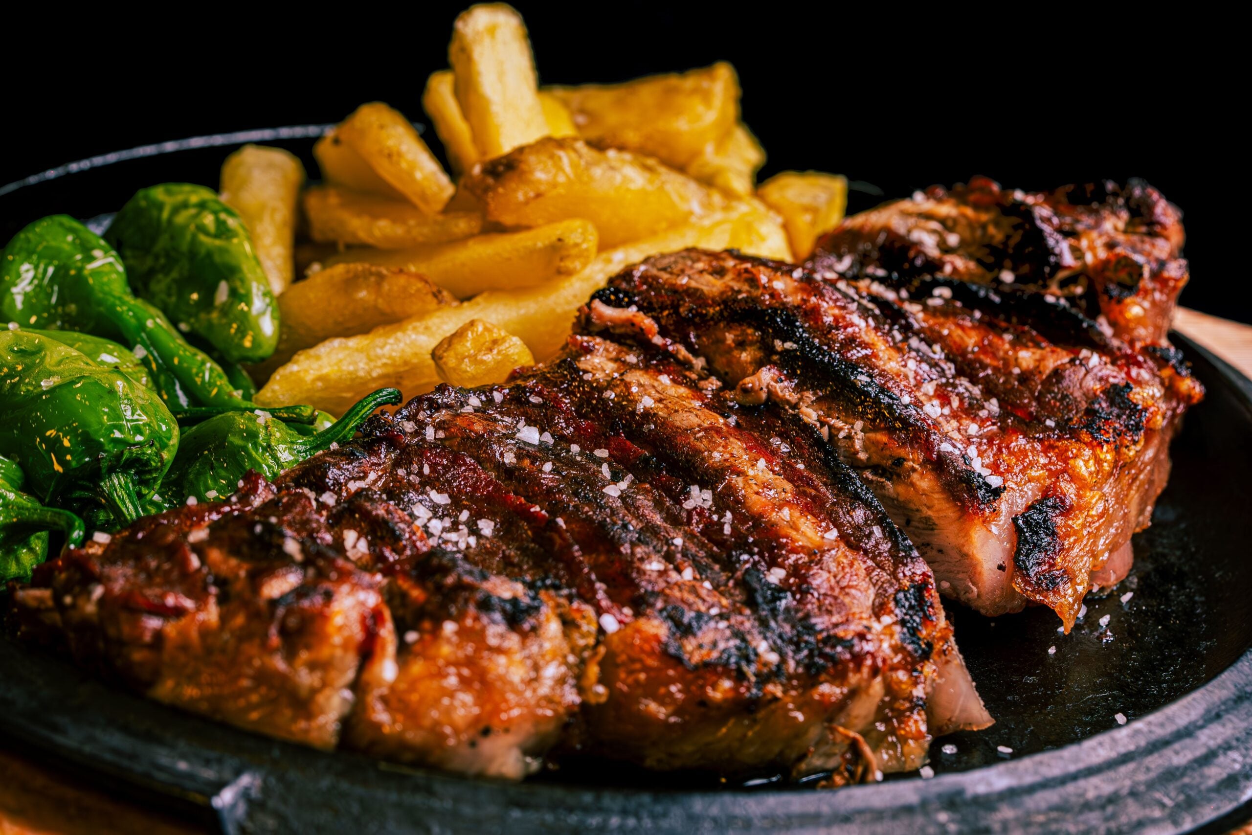 A close-up of a grilled steak sliced and topped with coarse salt, served with golden fries and green peppers on a black plate against a dark background.