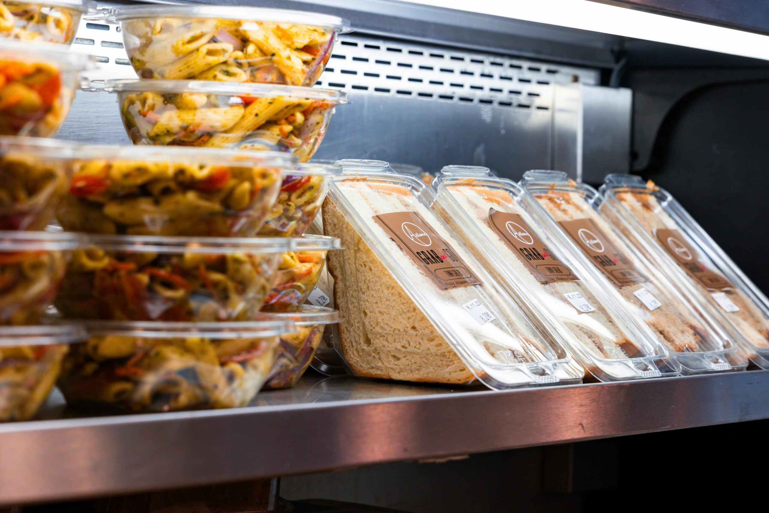 Plastic containers of pasta salad are stacked next to packaged sandwiches on a refrigerated shelf in a store or café display.