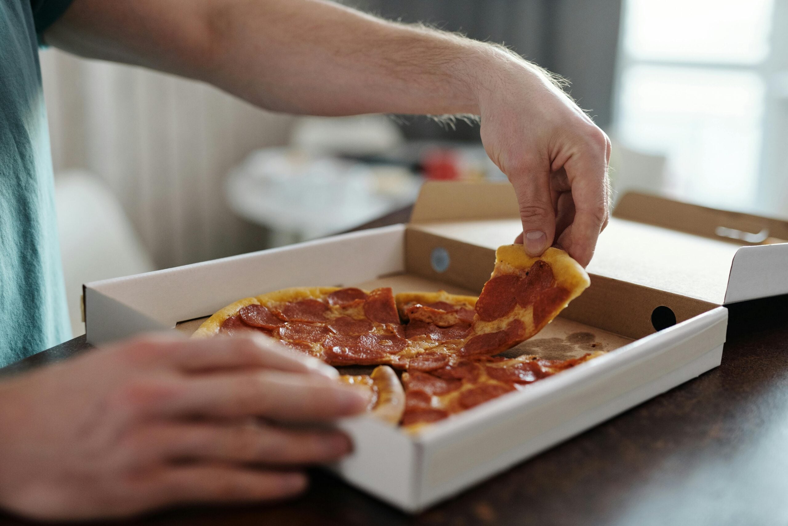 A person takes a slice of pepperoni pizza from an open pizza box on a dark table, with another hand steadying the box.