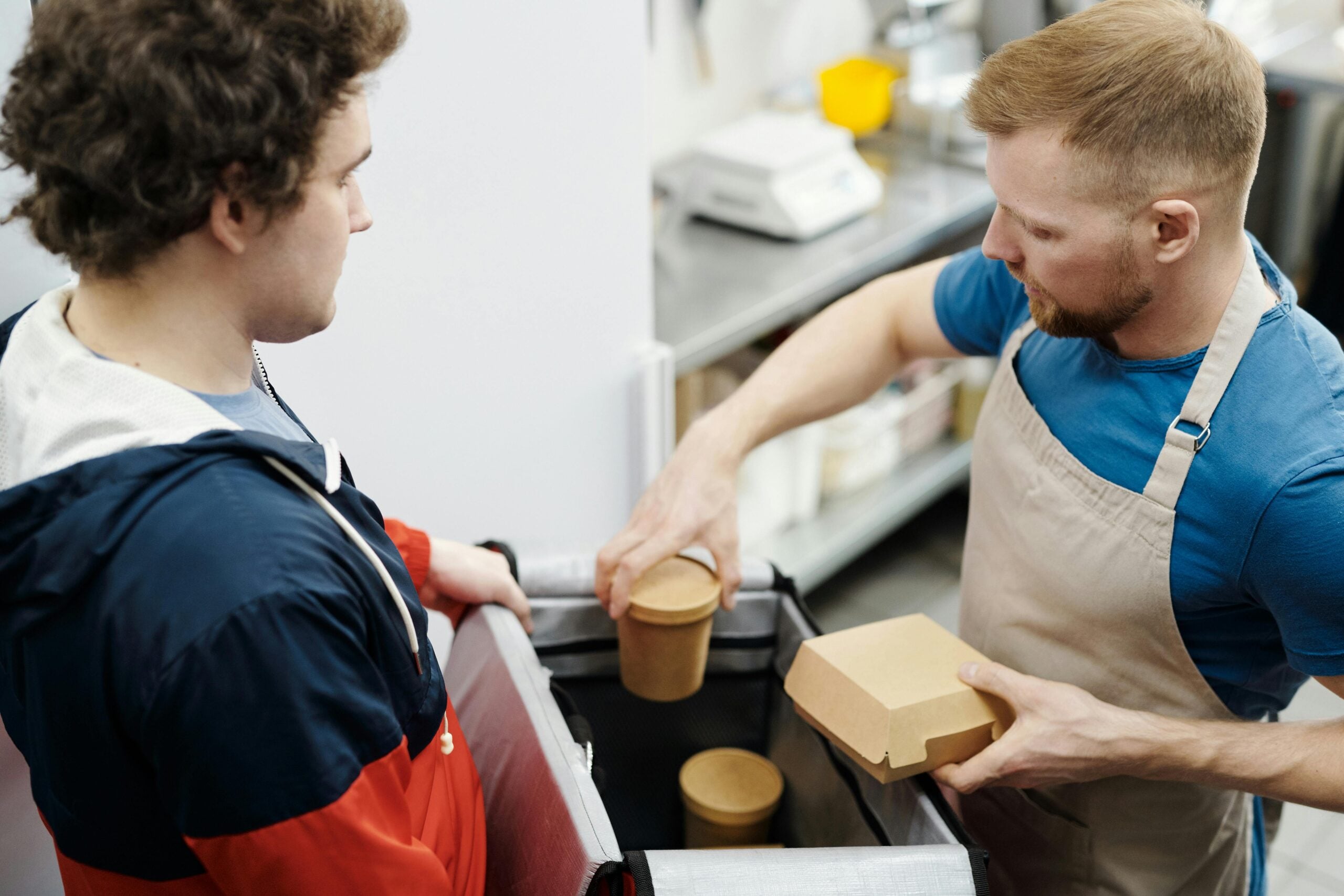 A man wearing an apron hands a cardboard food container and a brown paper cup to another man dressed in a red and navy jacket, likely at a food counter in a kitchen or cafe setting.