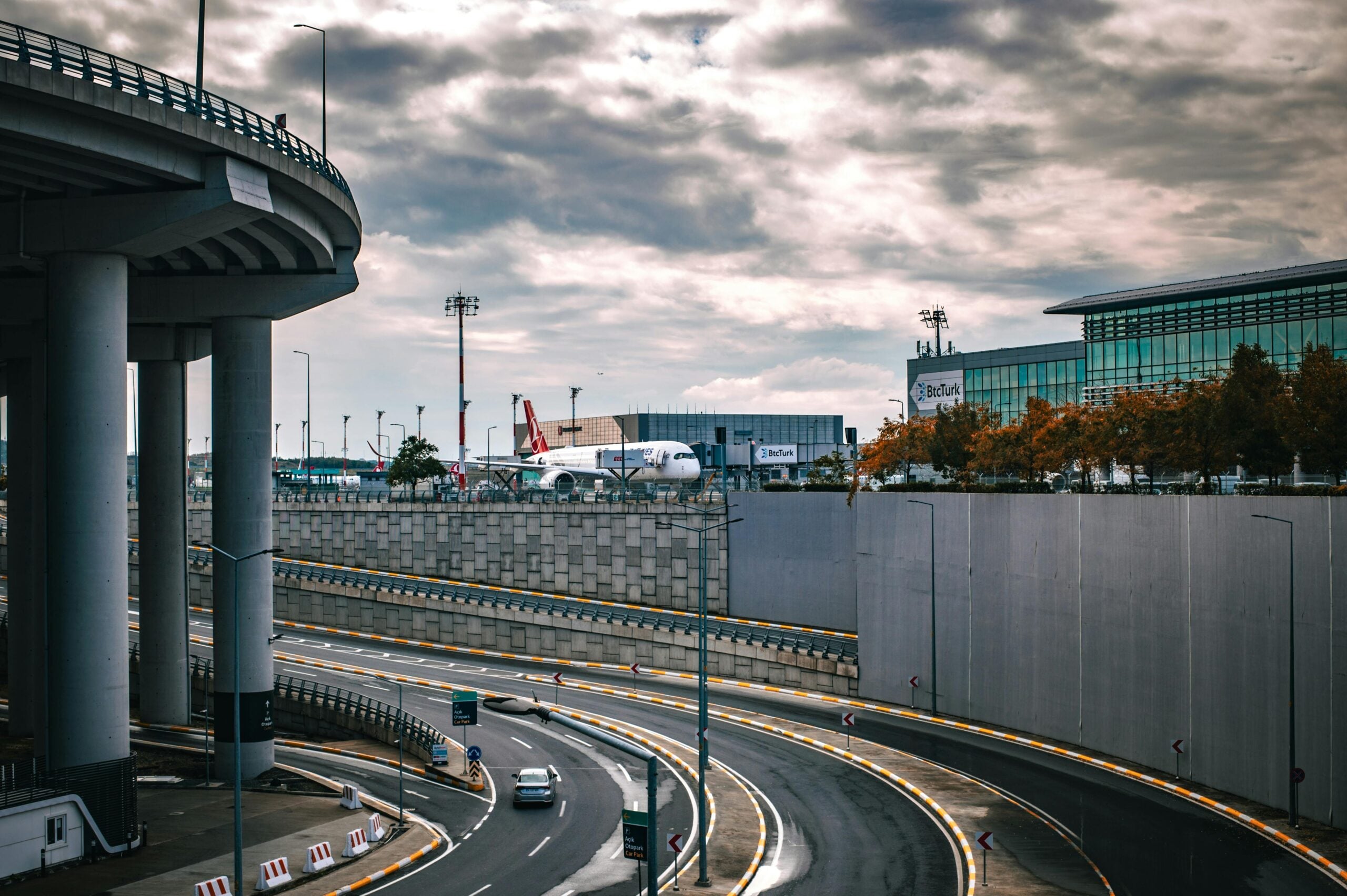 Curved roads lead to and from an airport; a large plane is visible near modern glass buildings under a cloudy sky. Elevated highway structures frame the scene.