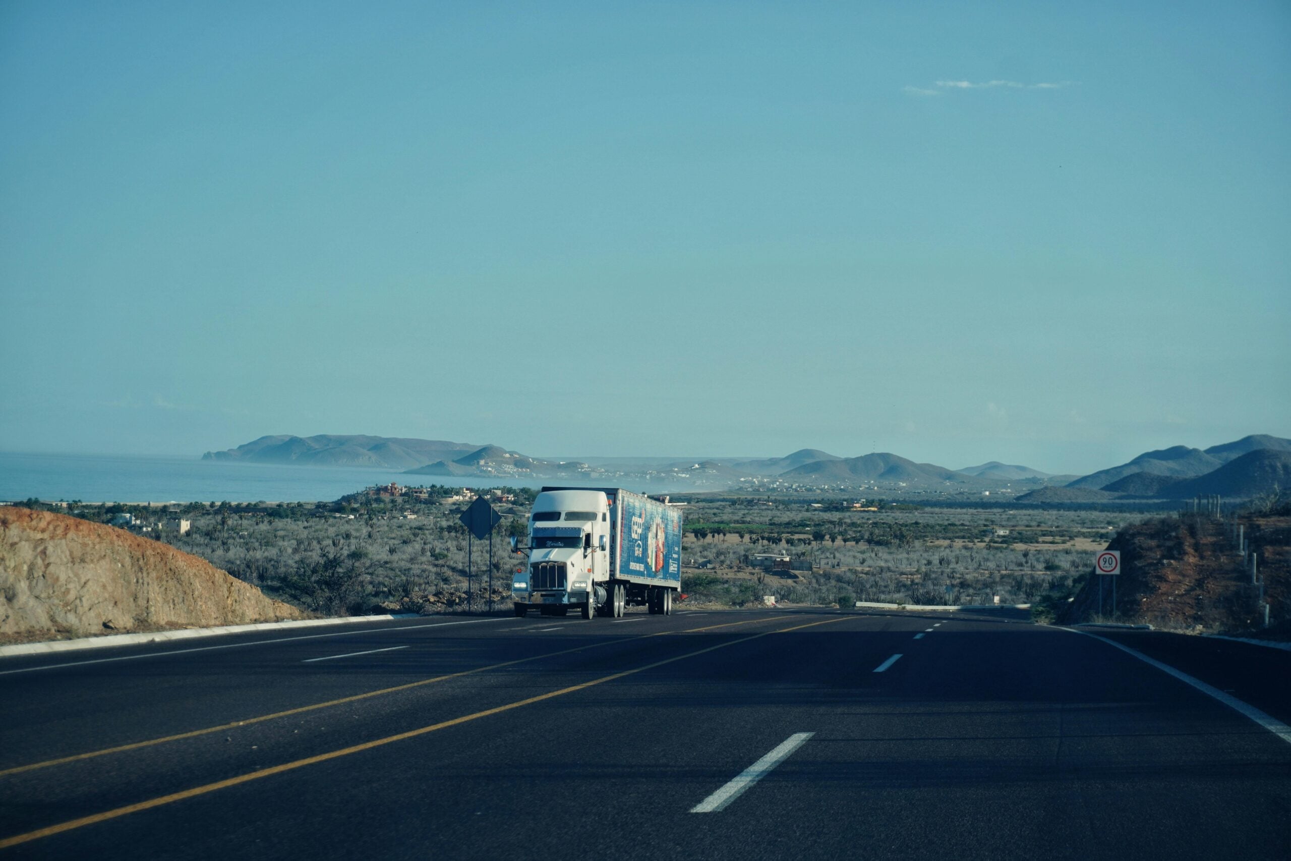 A white semi-truck drives along a wide, empty highway with mountains, greenery, and a glimpse of the ocean in the background under a clear blue sky.