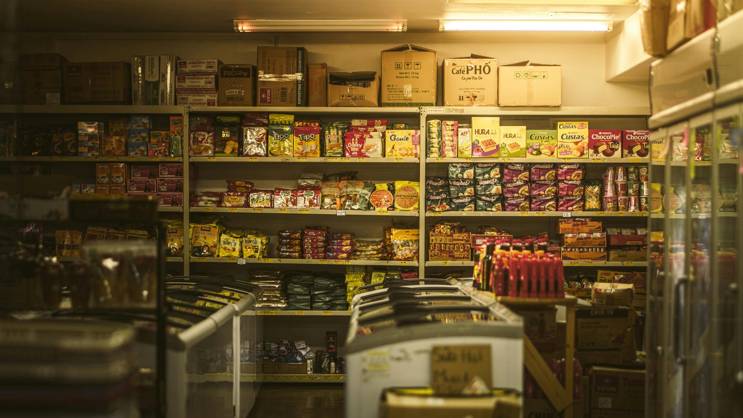A small grocery store interior with shelves stocked with colorful packaged food items, boxes on the top shelf, and coolers lining the aisles. The lighting is warm and the space appears tidy and organized.