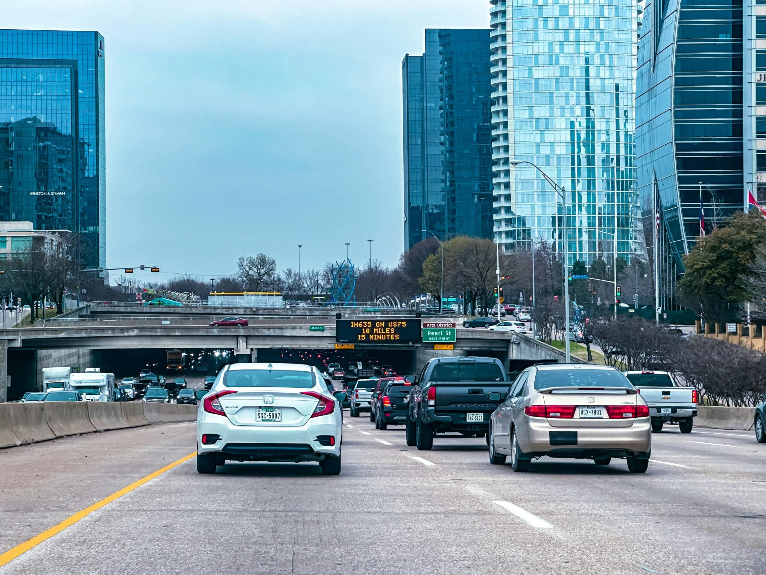 Cars drive on a busy city highway with tall modern buildings on both sides. An overhead electronic sign displays traffic information ahead, and trees line the roads under a cloudy sky.