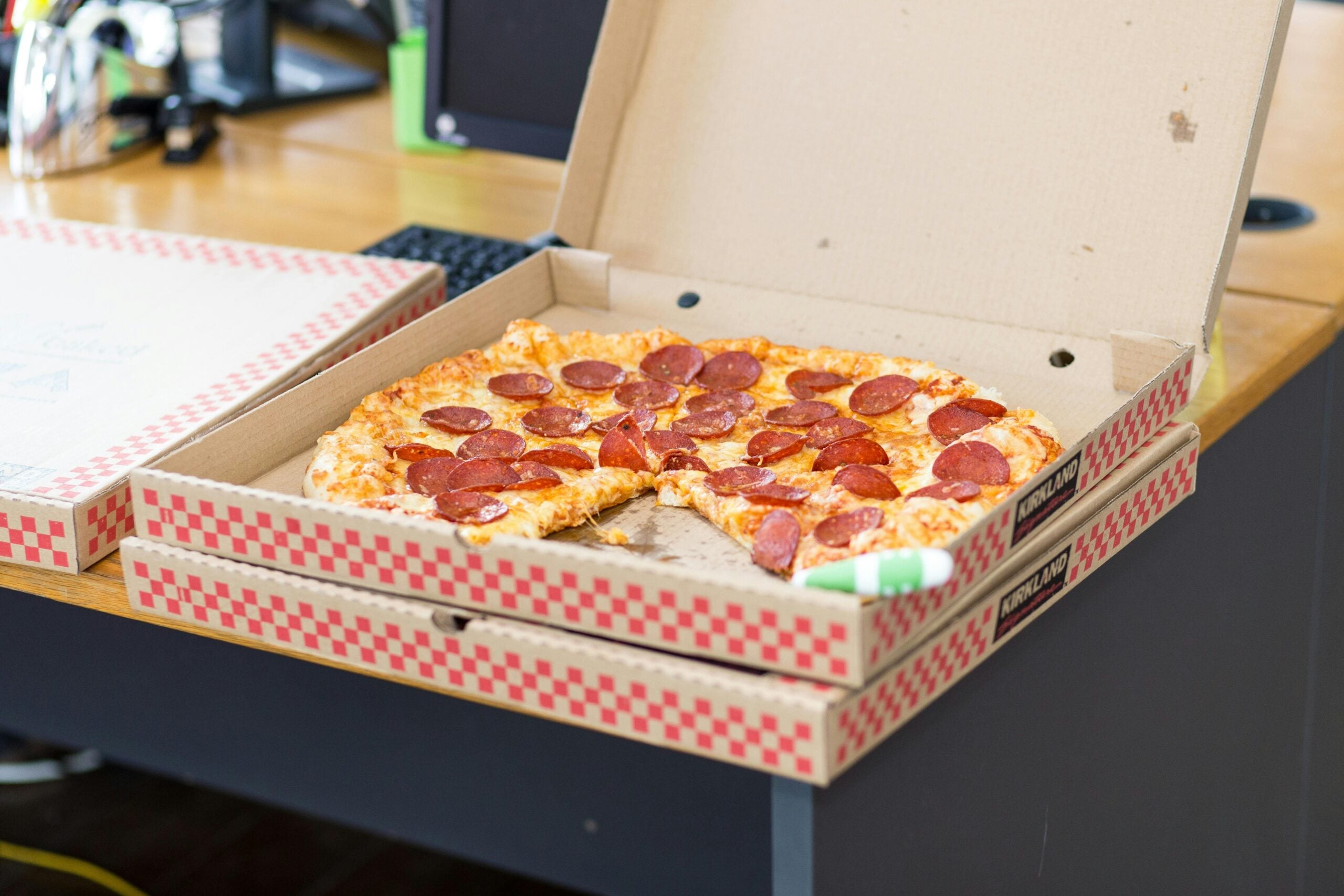 An open pizza box on a desk reveals a pepperoni pizza with several slices missing. Another closed pizza box is stacked underneath. Office supplies and a computer keyboard are visible in the background.