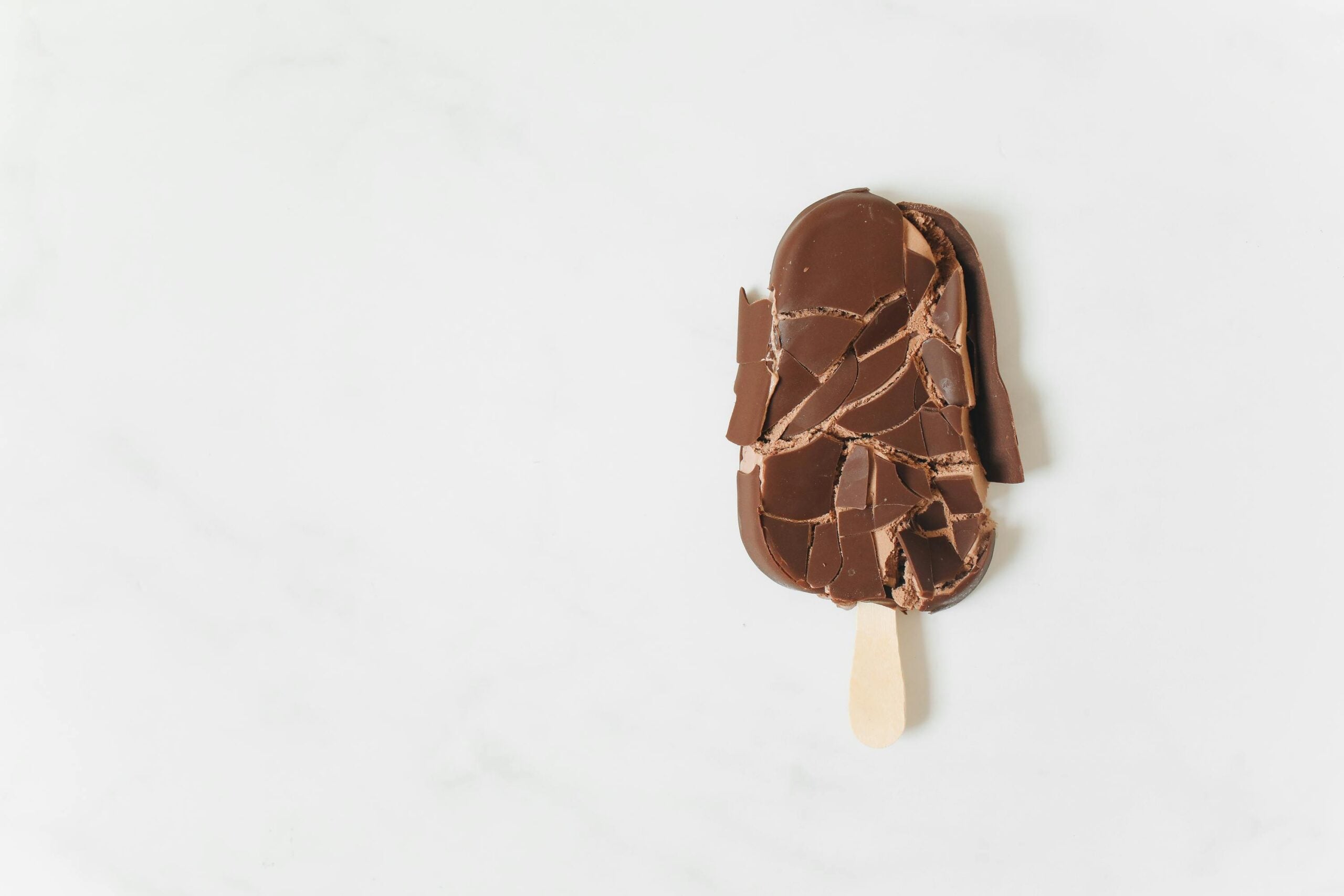 A chocolate-covered ice cream bar on a wooden stick lies on a white surface. The chocolate coating is broken into several pieces, revealing the ice cream beneath.